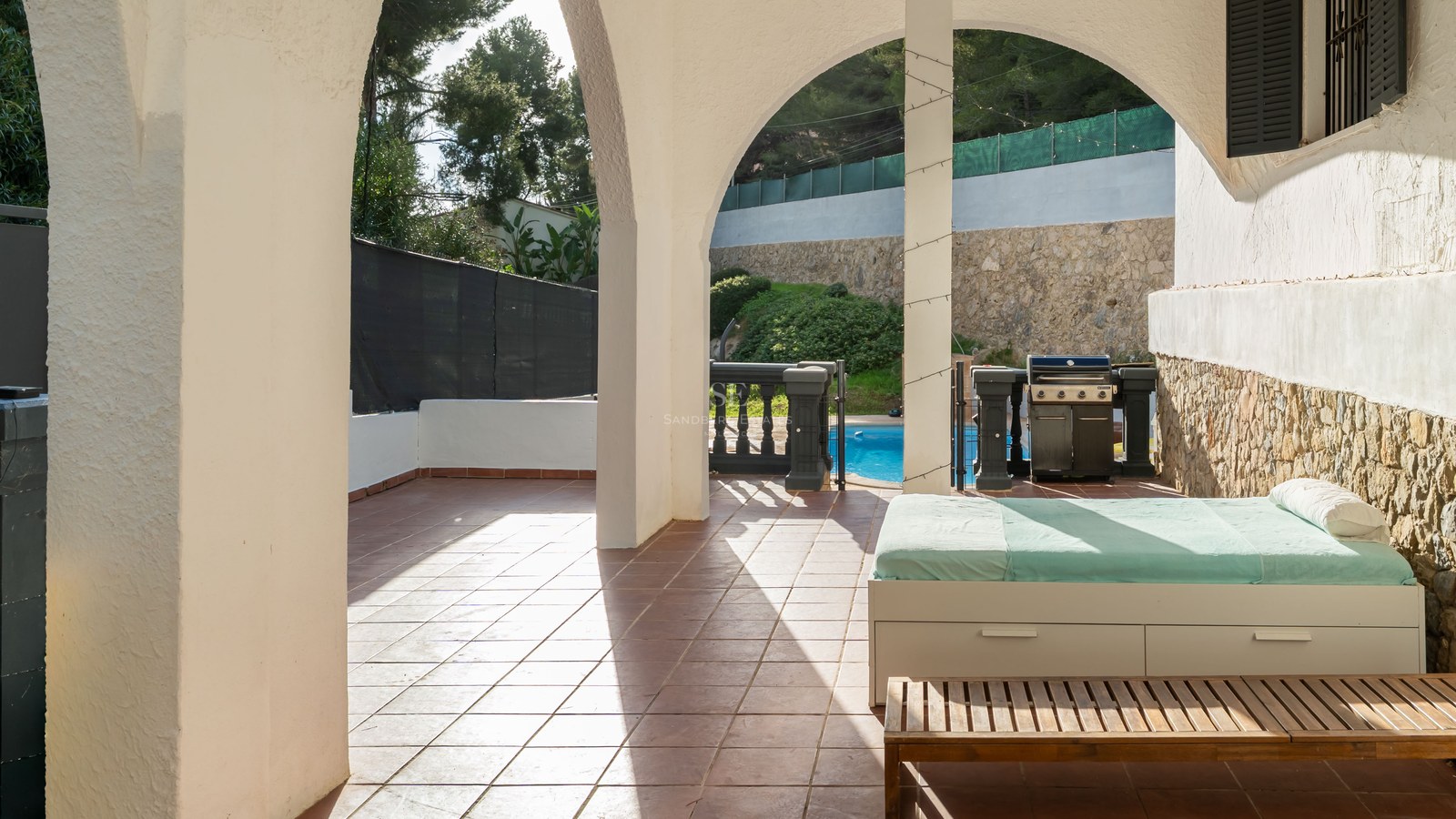Covered terrace with white arches, a daybed, BBQ grill, and view of a sunny swimming pool.