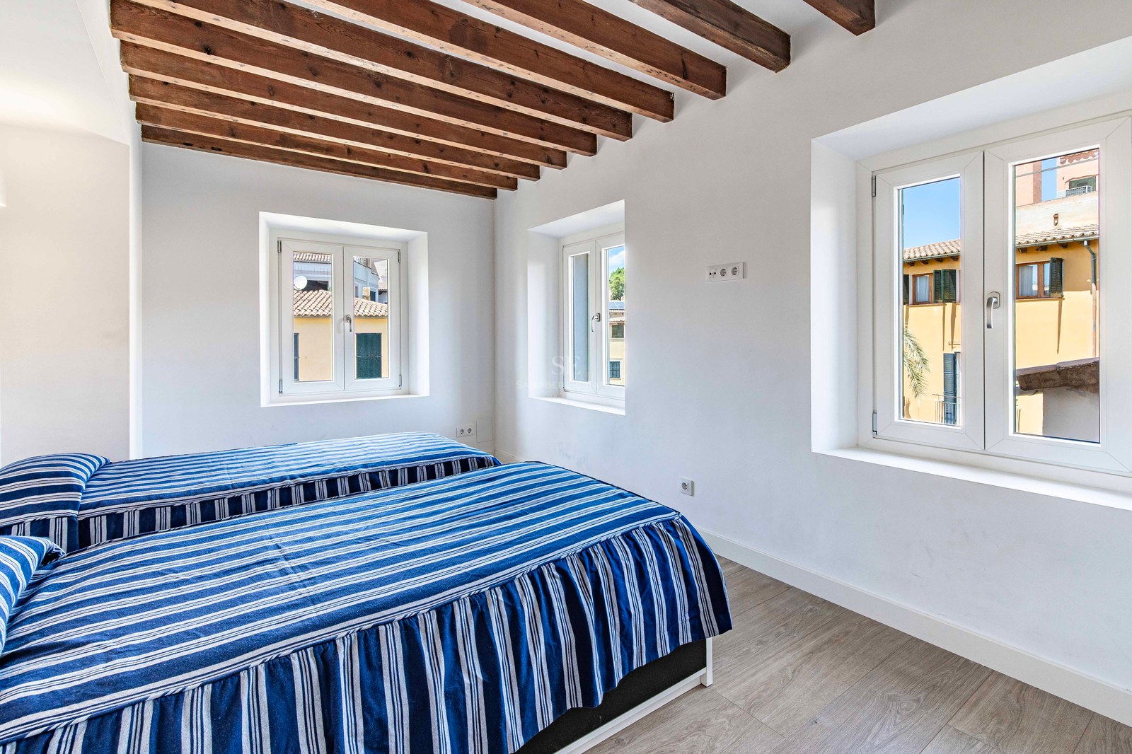 Bright bedroom featuring two twin beds, exposed dark wood ceiling beams, and views of historic town buildings.