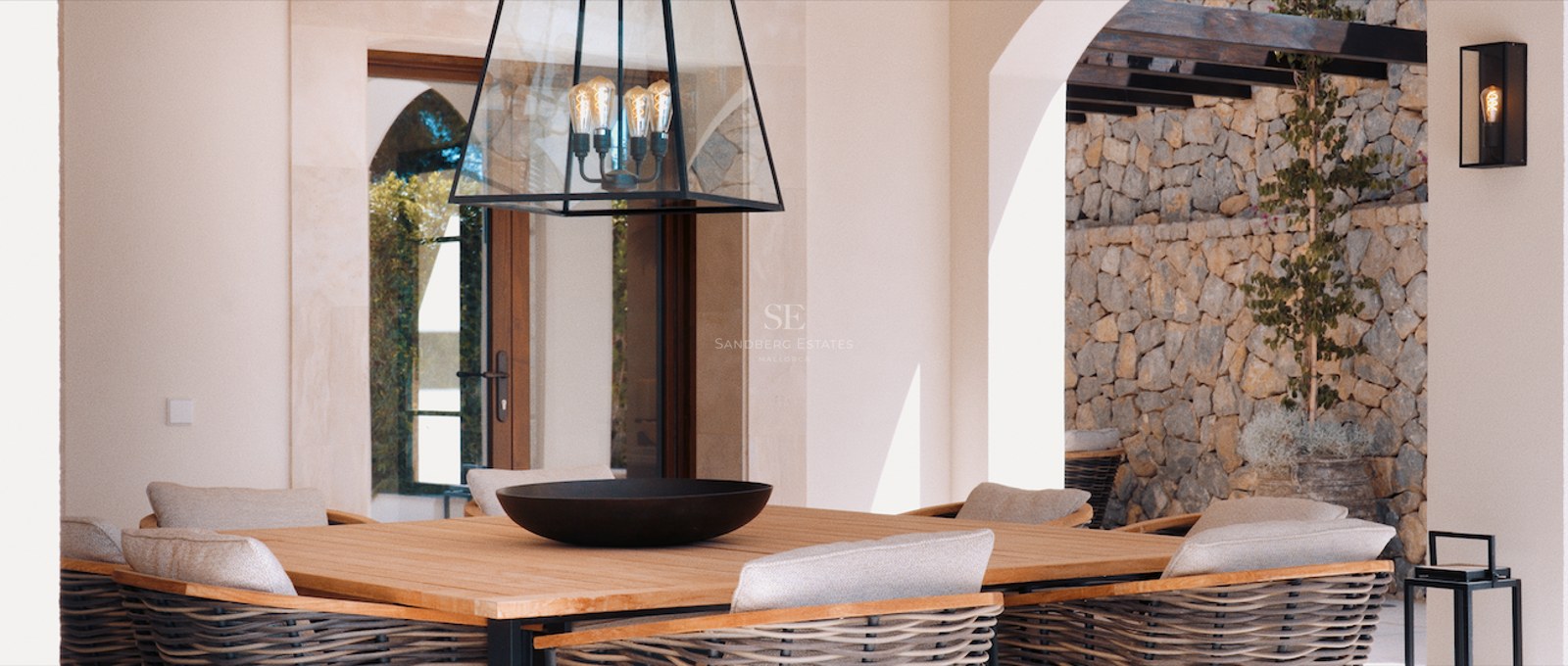 Wooden outdoor dining table with woven chairs on a luxury terrace featuring a stone wall and designer lighting.