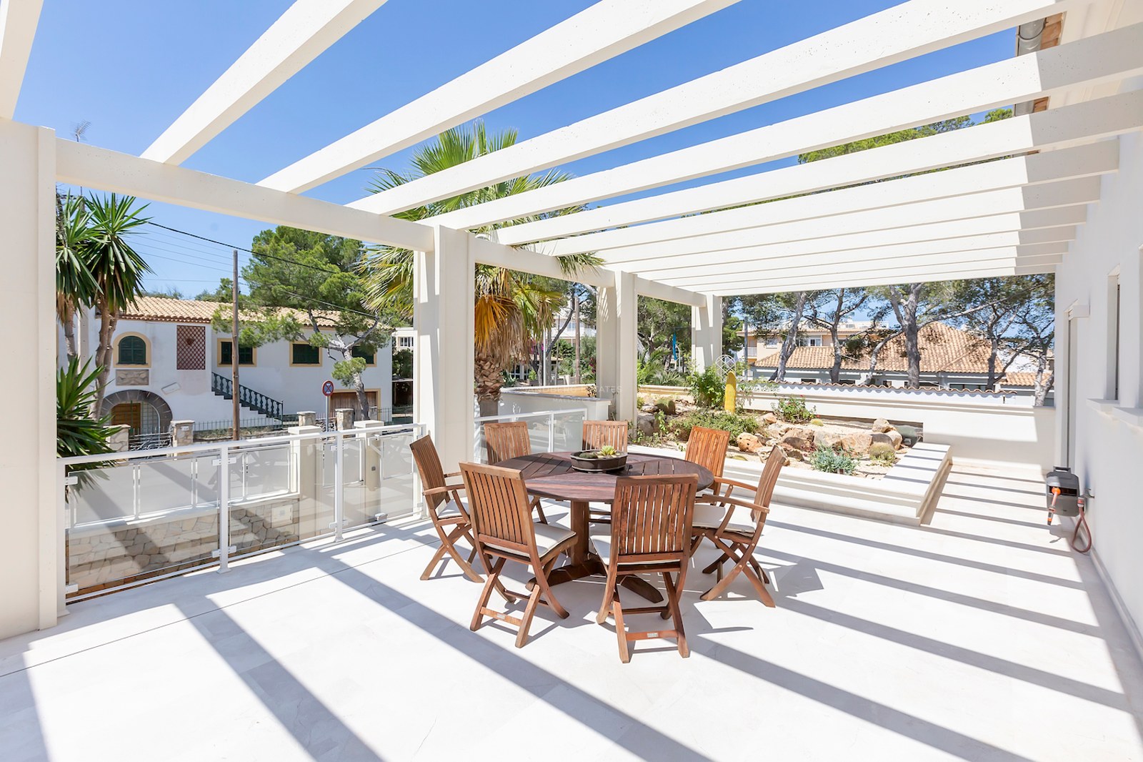 Sunny terrace featuring a white architectural pergola, round wooden dining set, and palm trees under a clear blue sky.