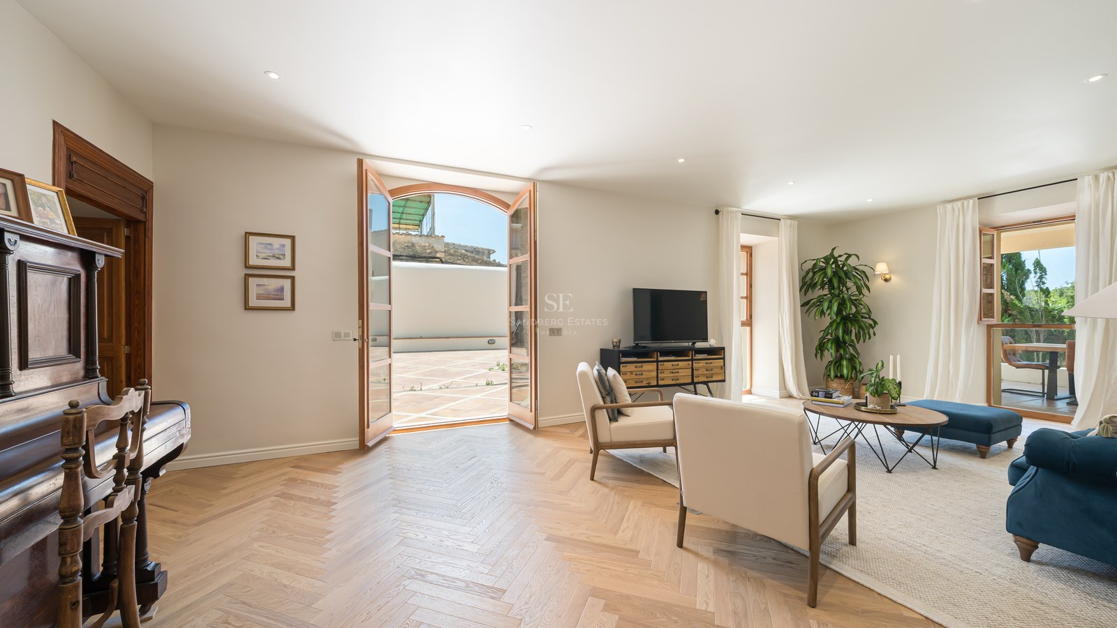 Bright living room featuring herringbone oak floors, cream armchairs, and glass doors opening to a sunlit terrace.