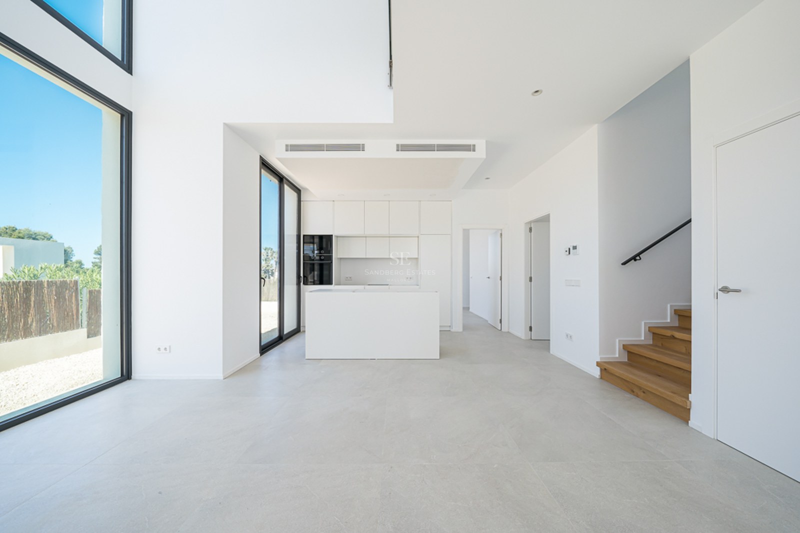 Cuisine blanche minimaliste avec îlot, escalier en bois et grandes baies vitrées du sol au plafond.