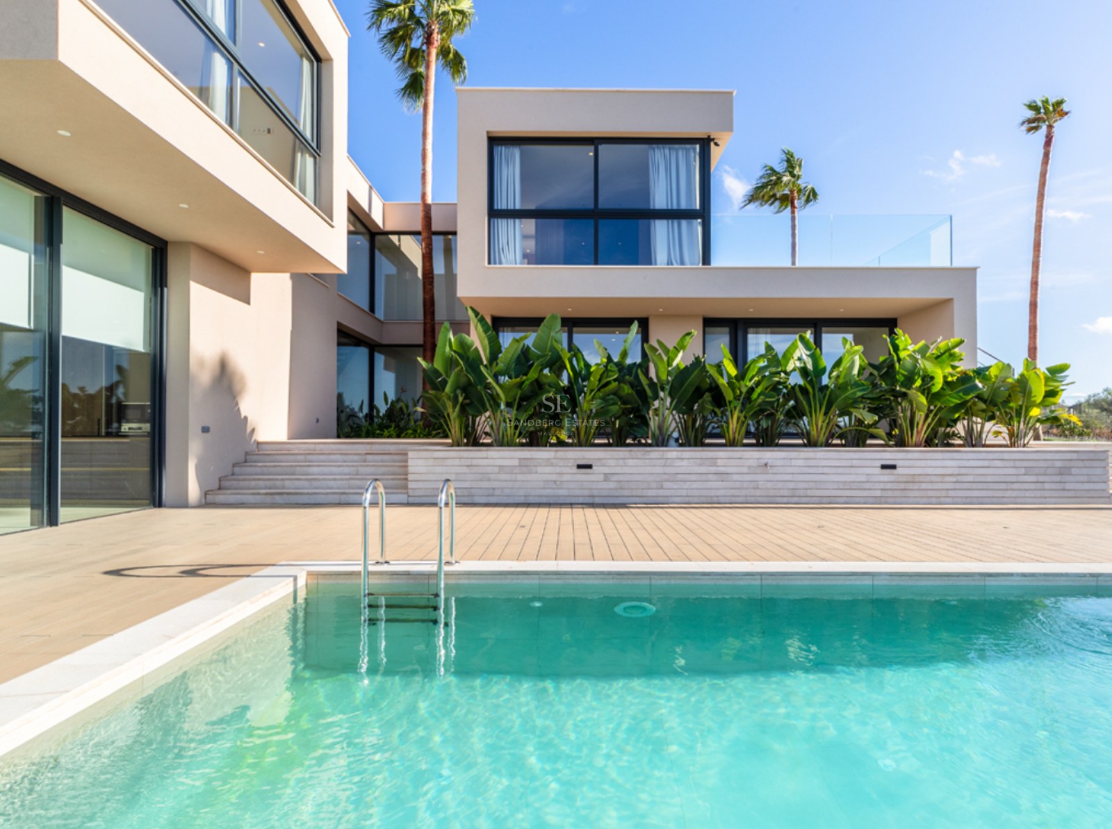 Modern beige villa med stora fönsterpartier mot en kristallklar pool och palmer under blå himmel.
