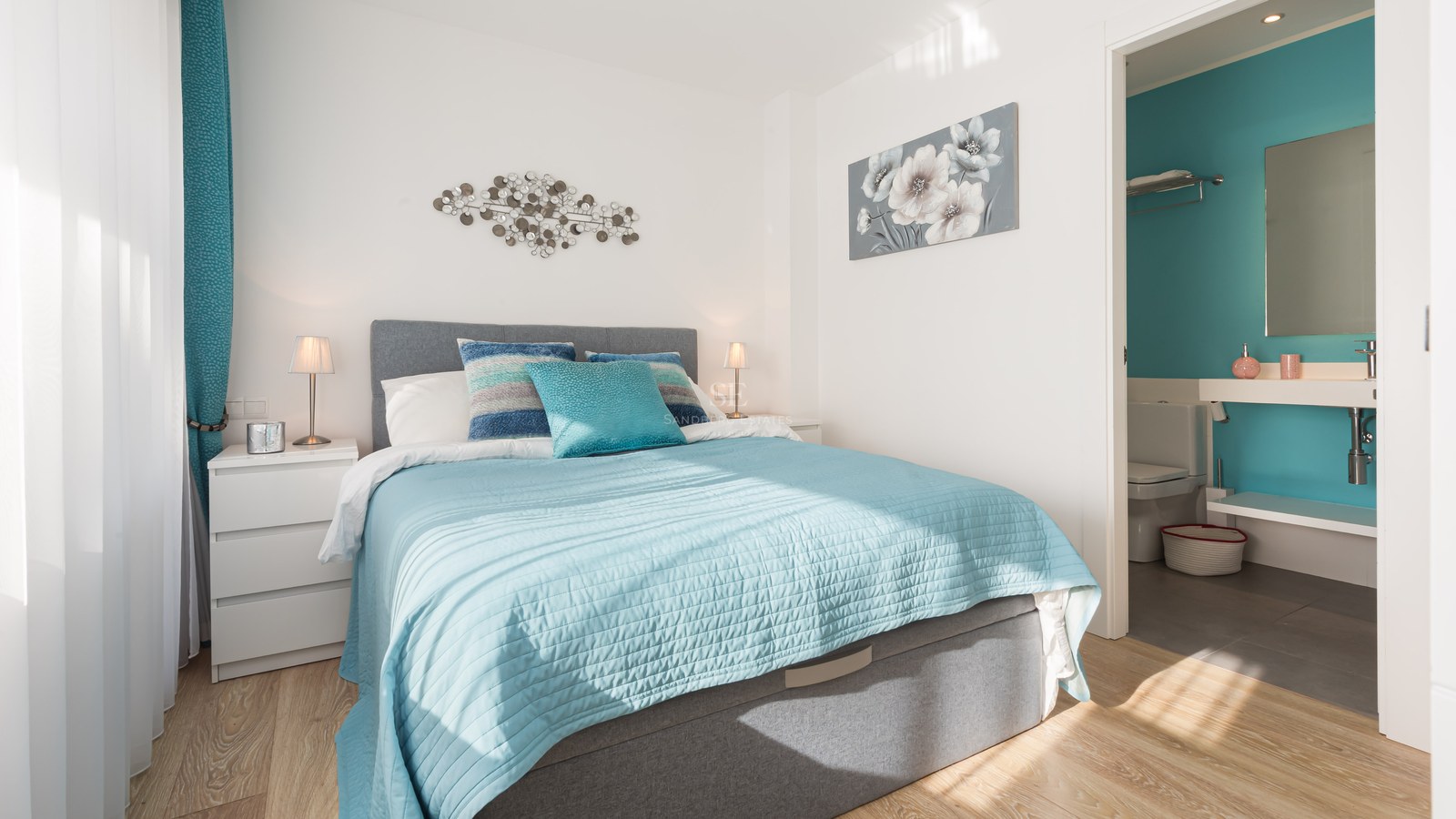 Bright bedroom featuring turquoise bedding, a grey upholstered headboard, and an open doorway to a matching ensuite bathroom.