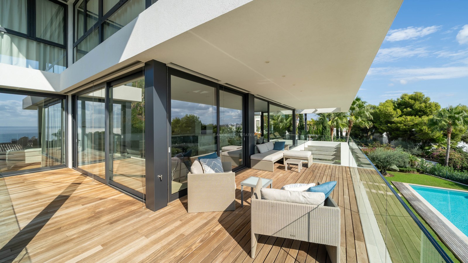 Terrasse spacieuse en bois avec mobilier lounge et garde-corps en verre surplombant une piscine et un jardin.