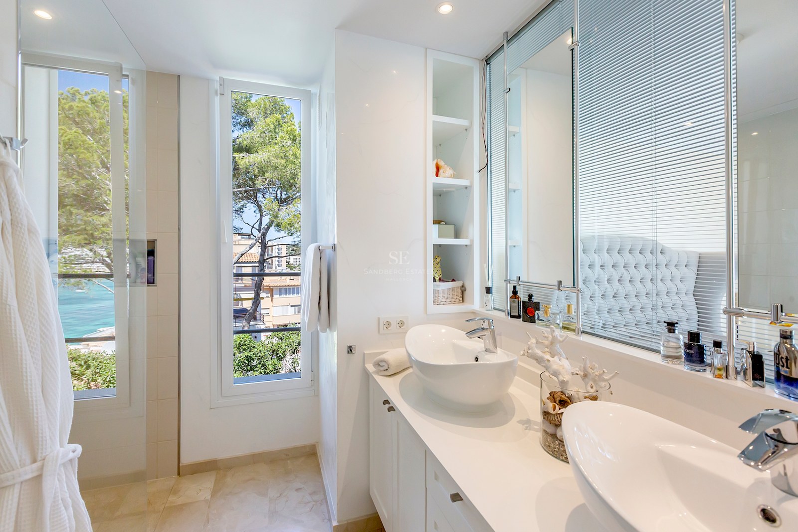 Bright bathroom featuring double white vessel sinks, a large mirror, and a window overlooking Mediterranean trees and the sea.