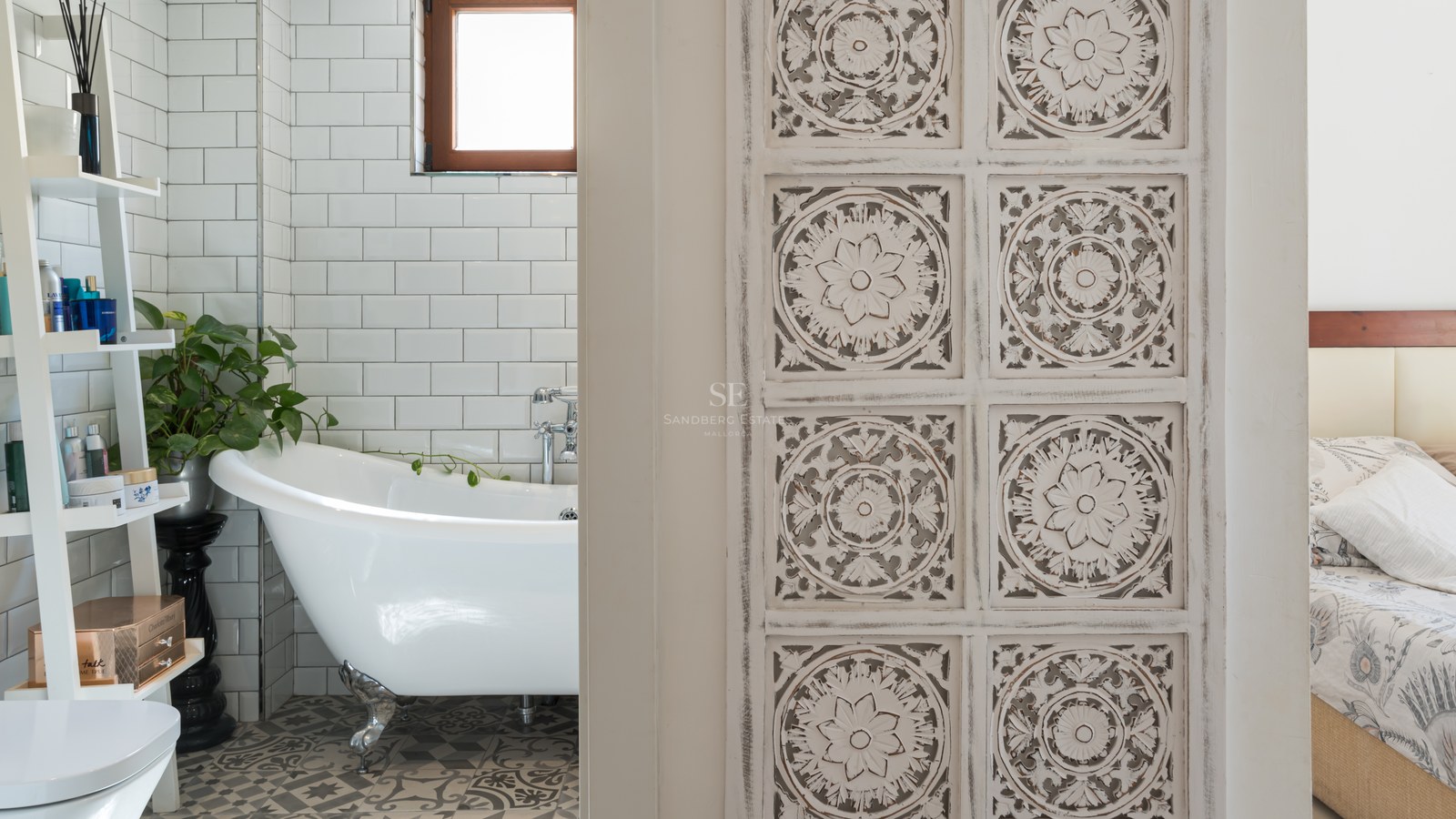 Un baño con bañera blanca de patas, azulejos tipo metro y suelo hidráulico, visto a través de una puerta de madera tallada.