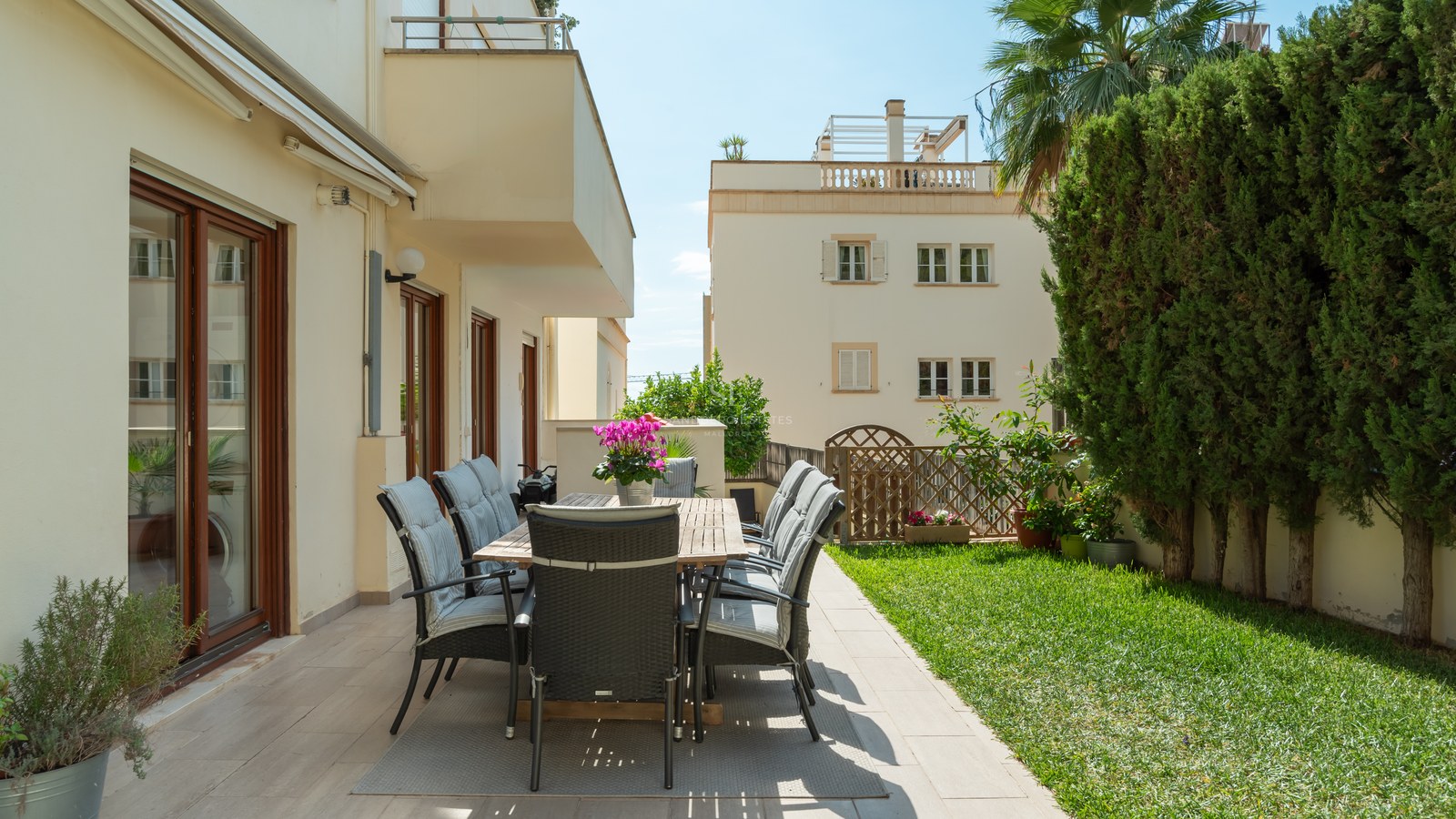 Comedor exterior en terraza de baldosas junto a jardín con césped y setos altos bajo luz solar directa.