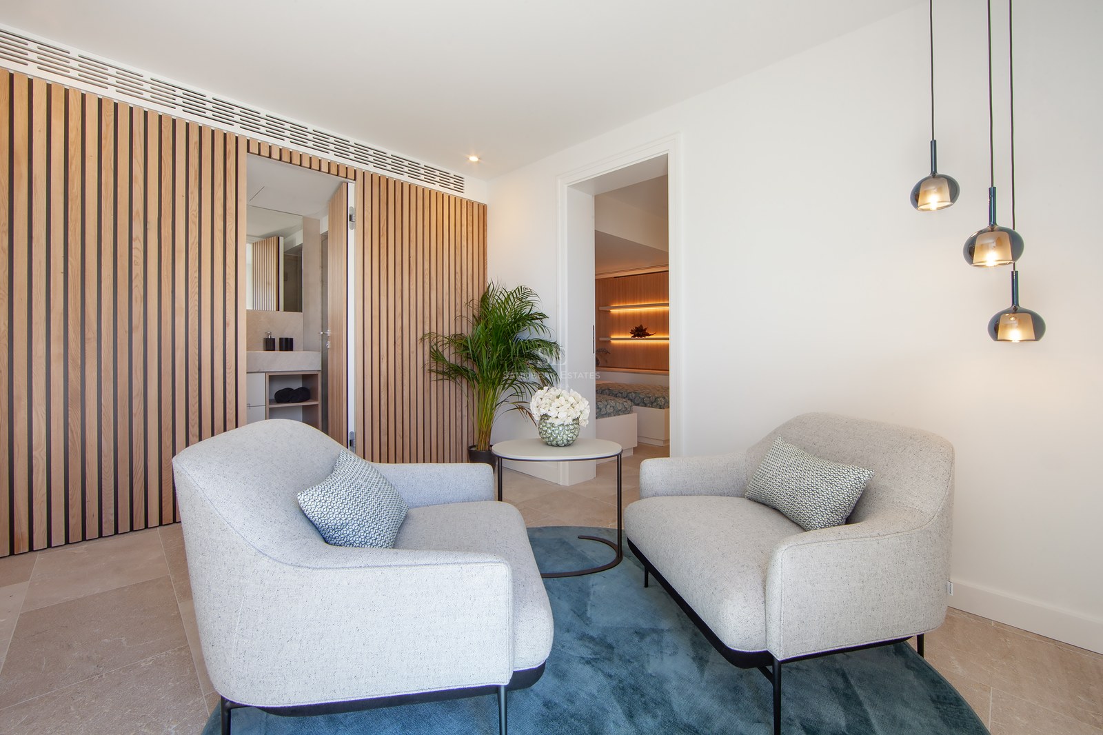 Master bedroom suite featuring two grey armchairs, blue rug, wooden slat wall, and view of the bedroom area.