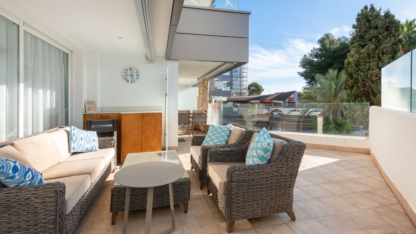 Spacious terrace with grey wicker sofas, blue accent cushions, and glass railings under a clear blue sky.