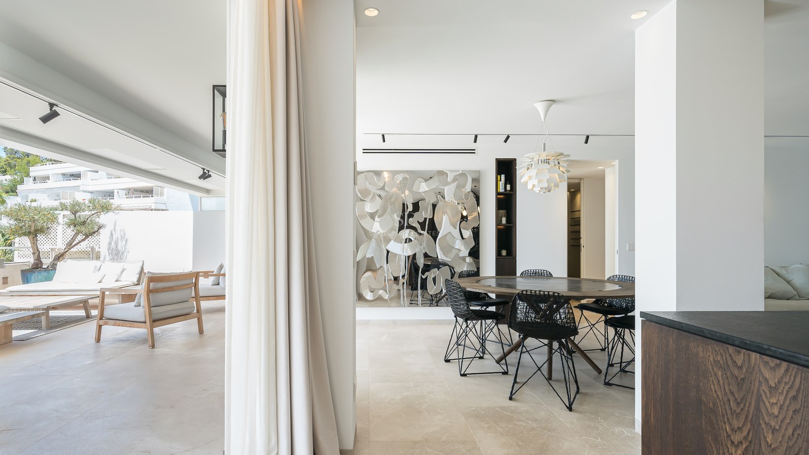 Salle à manger moderne avec table ronde en bois s'ouvrant sur une terrasse ensoleillée avec mobilier de salon.