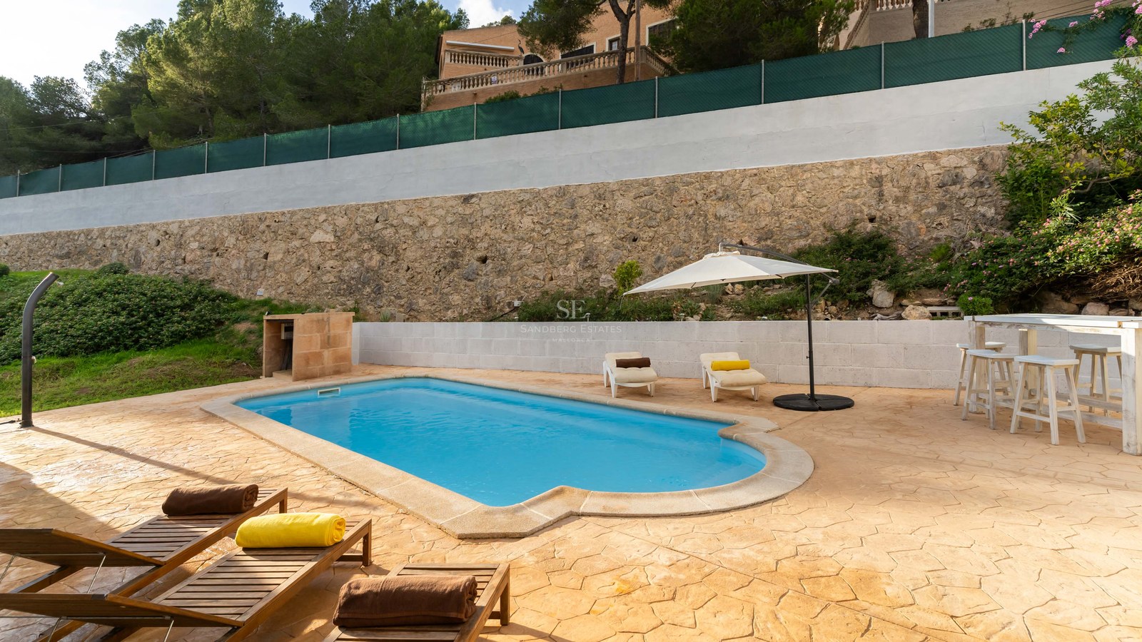 Outdoor pool with stone terrace, lounge chairs, white umbrella, and stone wall backdrop.