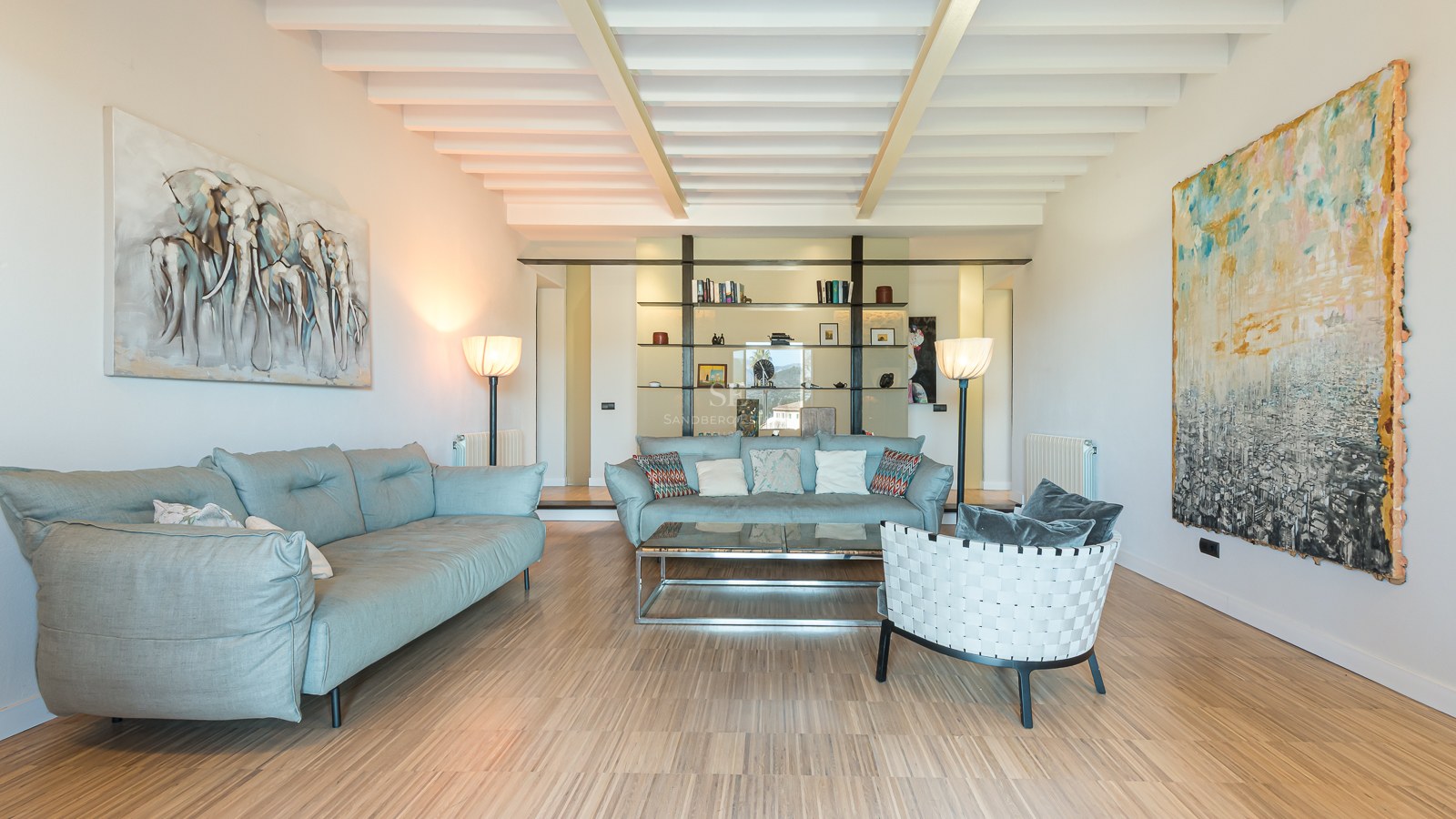 Spacious living room with light blue sofas, white beamed ceiling, wooden floors, and large abstract paintings.