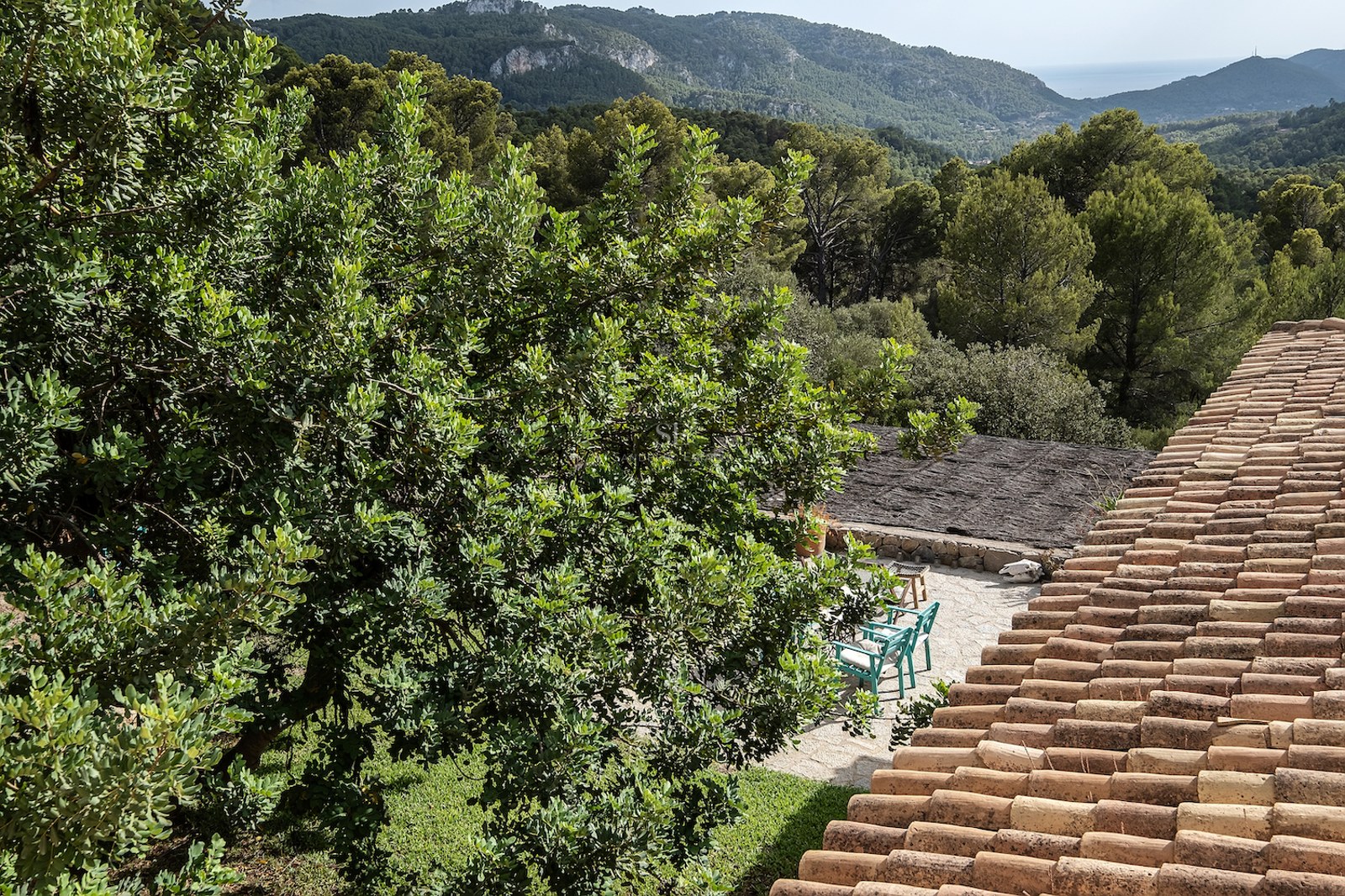 Toit en terre cuite surplombant un patio en pierre avec des chaises turquoise, entouré d'arbres et de montagnes.