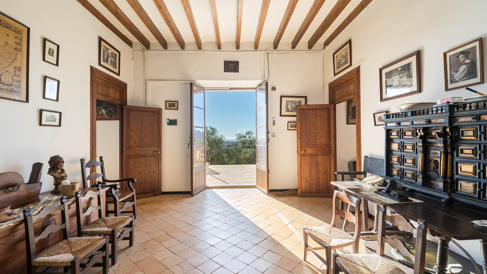 Vestíbulo tradicional con vigas de madera, suelo de terracota y puertas abiertas hacia una terraza soleada.