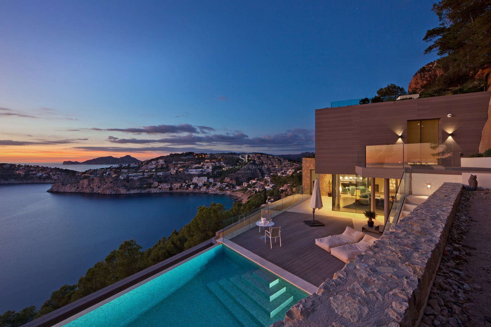 Modern villa terrace with infinity pool overlooking a bay at sunset with mountains in the background.
