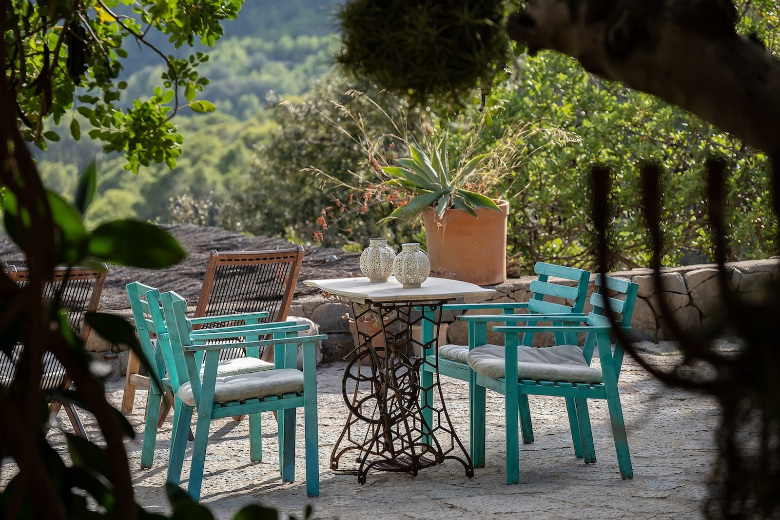 Table en marbre vintage et chaises en bois bleu canard sur un patio en pierre entouré de verdure méditerranéenne.