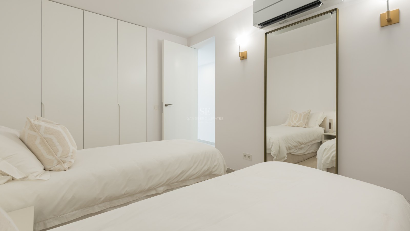 Bright minimalist bedroom featuring two twin beds, floor-to-ceiling white wardrobes, and a large gold-framed mirror.