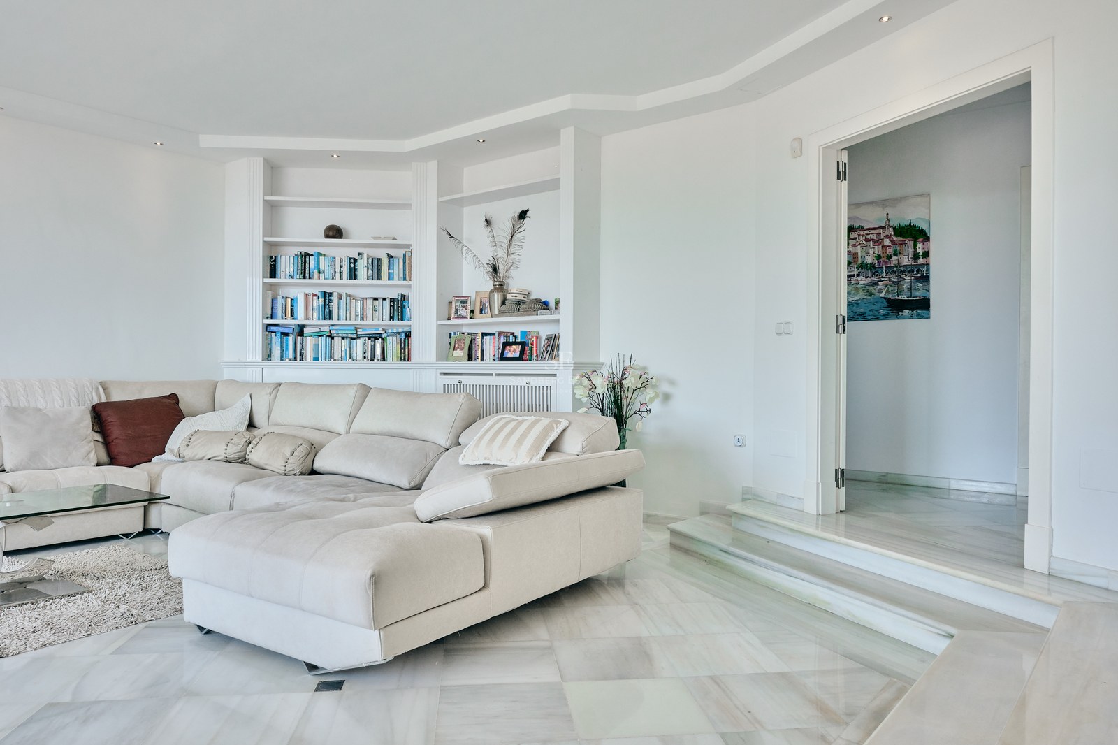 Contemporary living room with white marble floors, a large L-shaped cream sofa, and built-in white bookshelves.