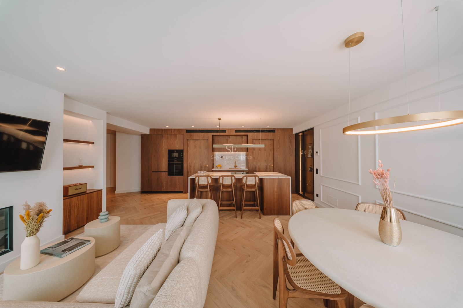 Grand salon luxueux avec canapé beige, parquet en chevrons et cuisine intégrée en bois naturel.