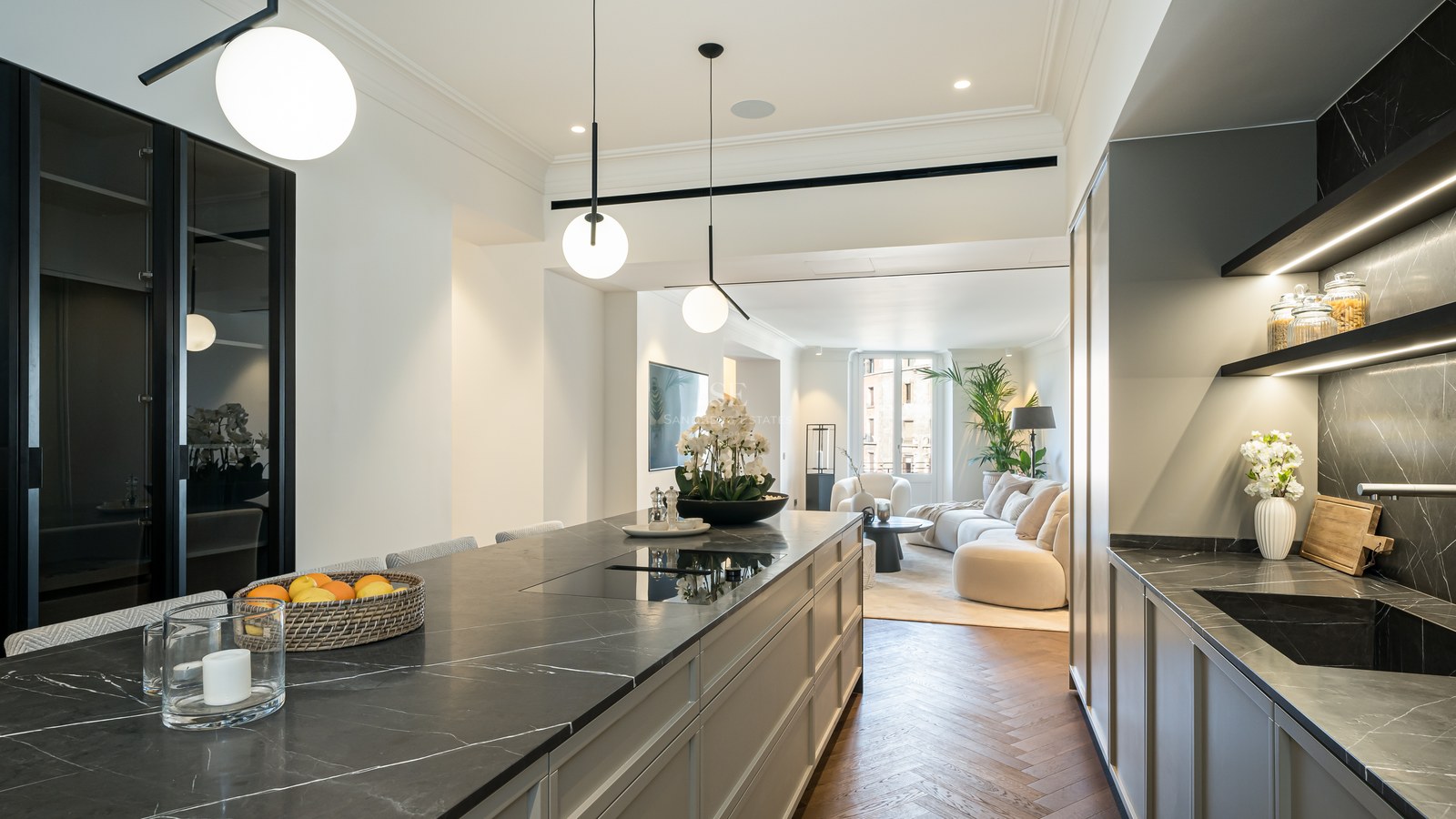 Cocina moderna con isla de mármol oscuro y suelos de espiga, conectada a un salón luminoso.