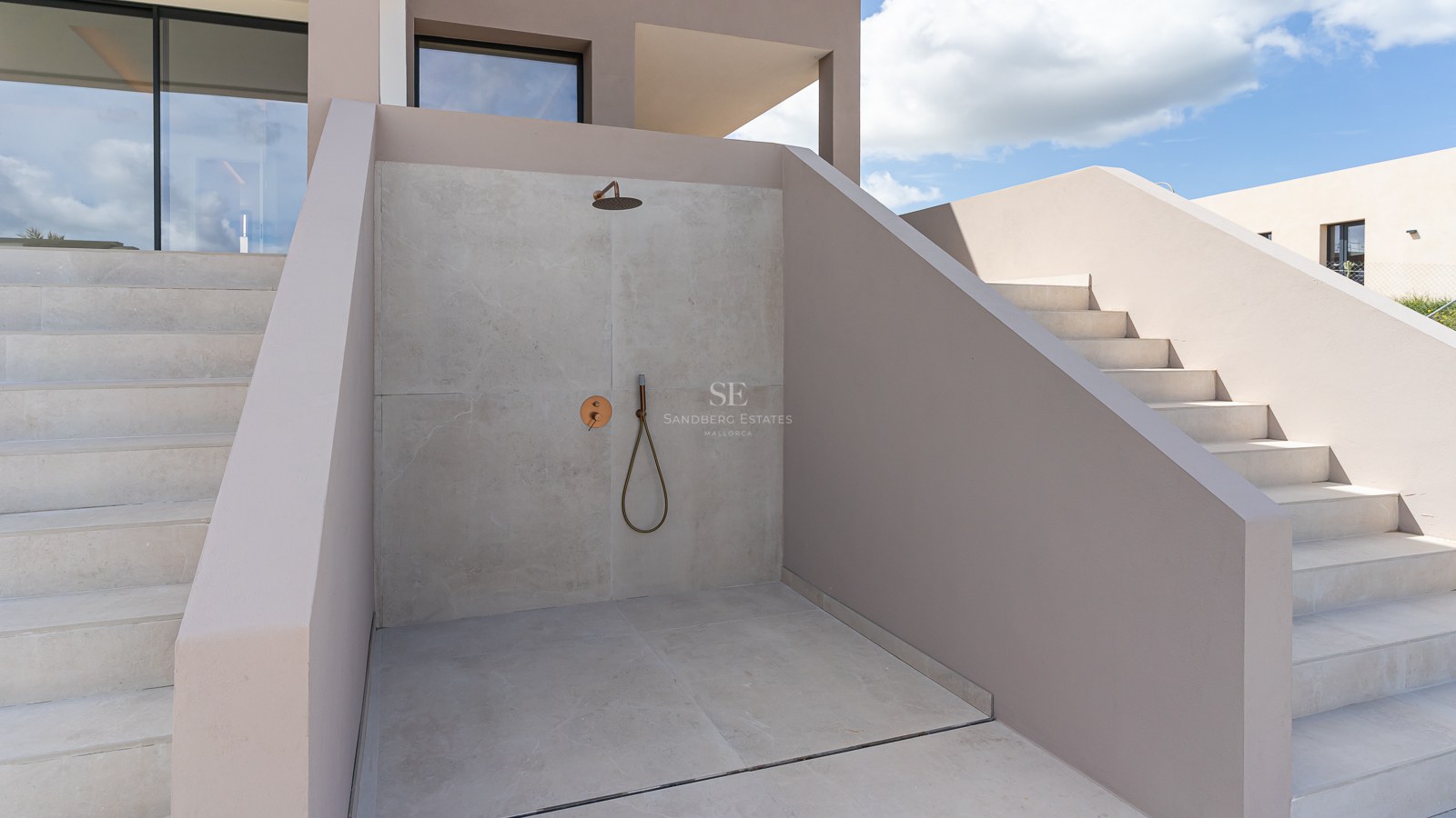 Minimalist outdoor shower with light gray microcement walls, brass fixtures, and a clear sky background.