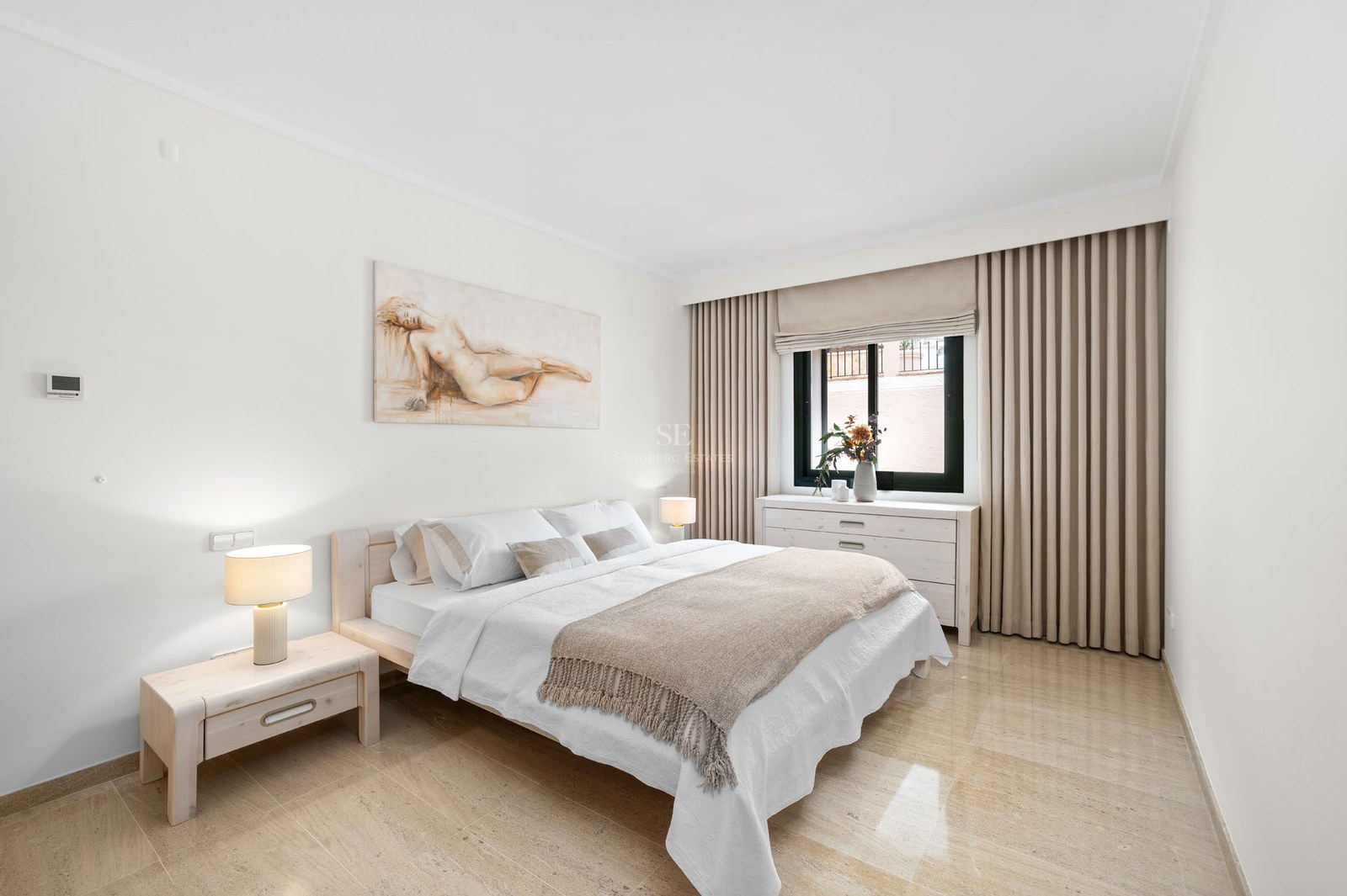 Chambre lumineuse avec lit en bois clair, sol en marbre, murs blancs et un grand tableau au-dessus du lit.