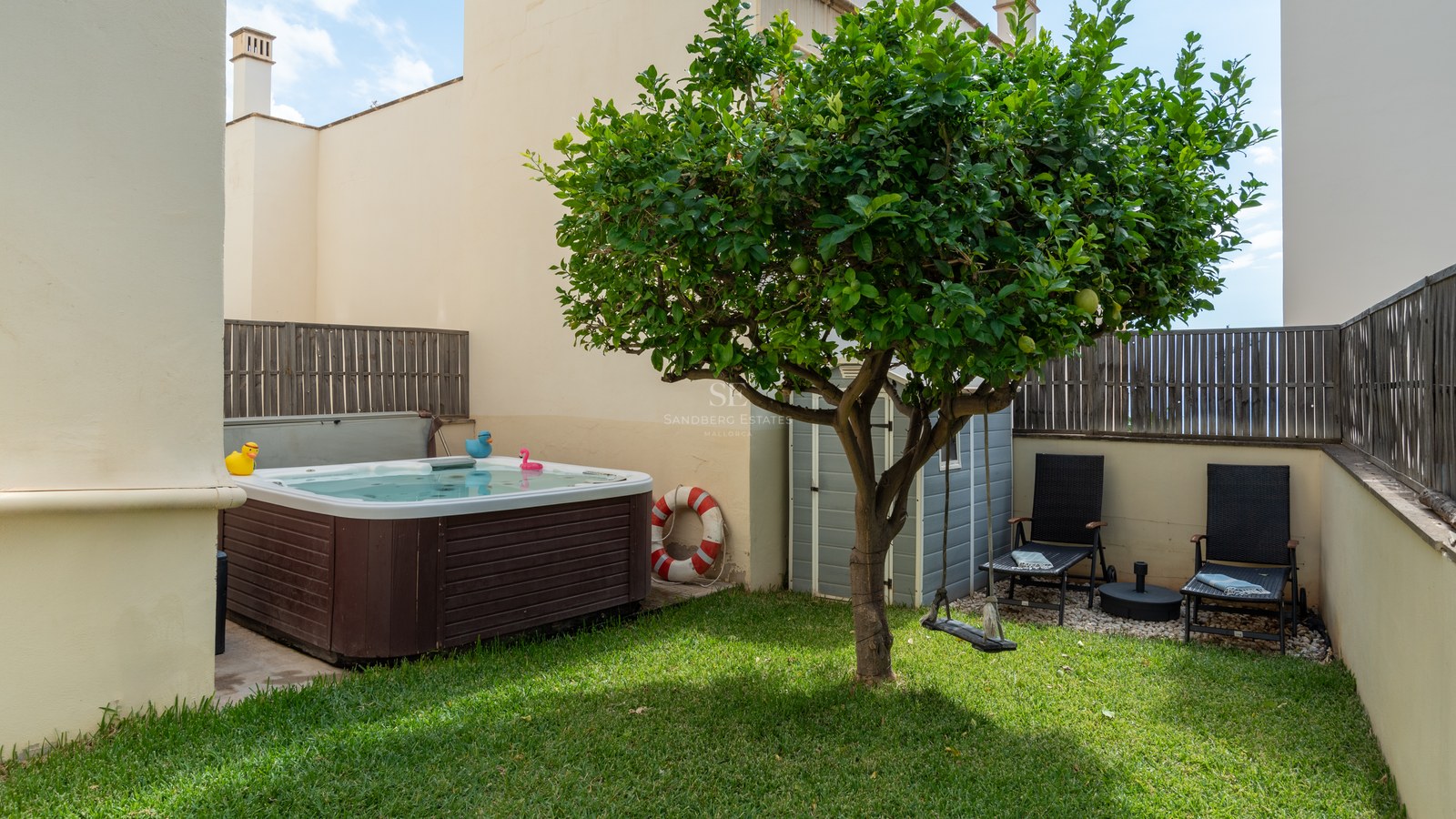 Pequeño jardín privado con césped, jacuzzi de madera, un limonero central y dos hamacas negras.