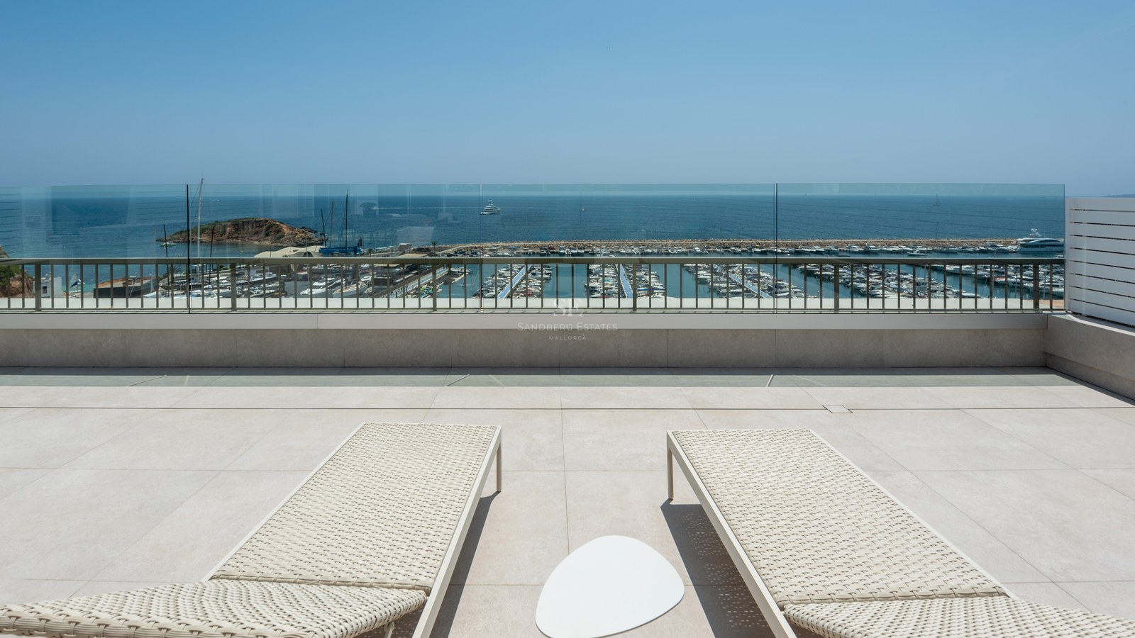 Two cream sun loungers on a stone terrace overlooking a yacht marina and the Mediterranean Sea under a clear sky.