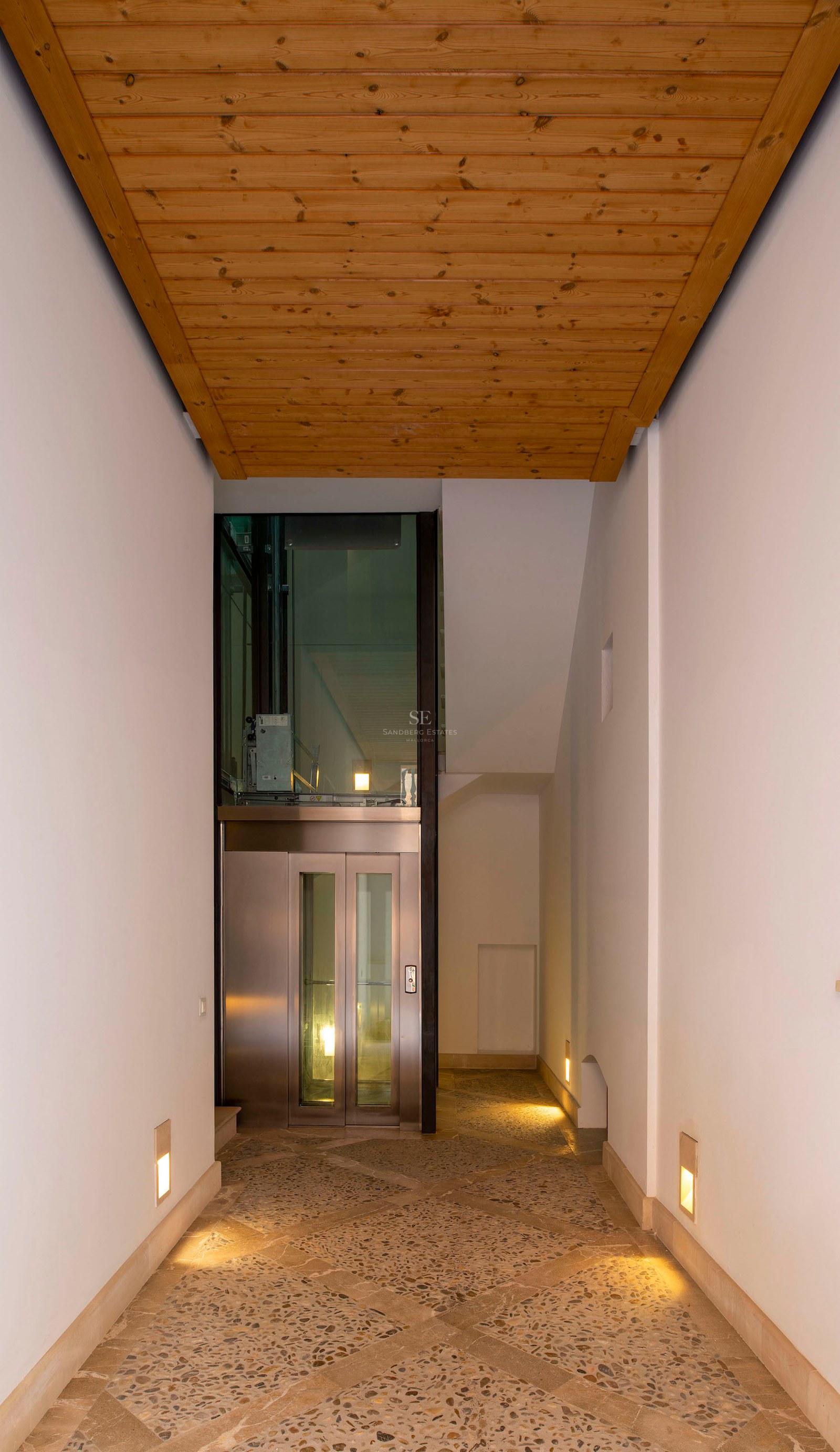 Modern hallway with stone mosaic flooring, wood ceiling, and a glass elevator at the end.