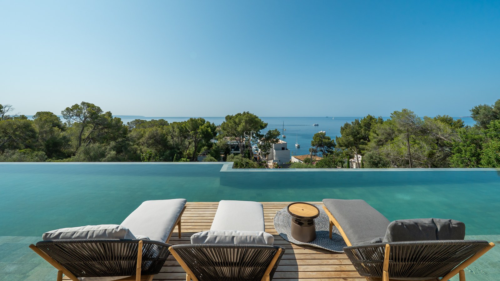 Three modern loungers on a wooden deck overlooking a large infinity pool and the sea with pine trees.