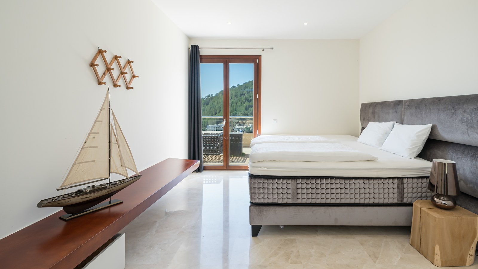 Dormitorio luminoso con suelos de mármol, cama de terciopelo gris y puerta de cristal con vistas a la montaña.