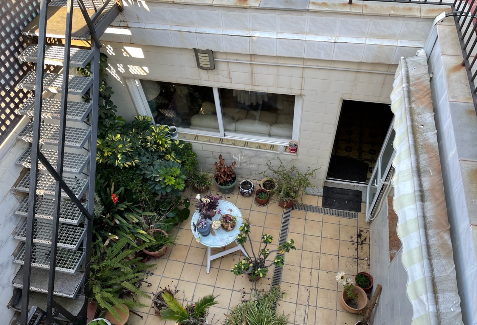 Vue d'en haut d'une terrasse carrelée avec escalier métallique, nombreuses plantes en pots et petite table ronde.