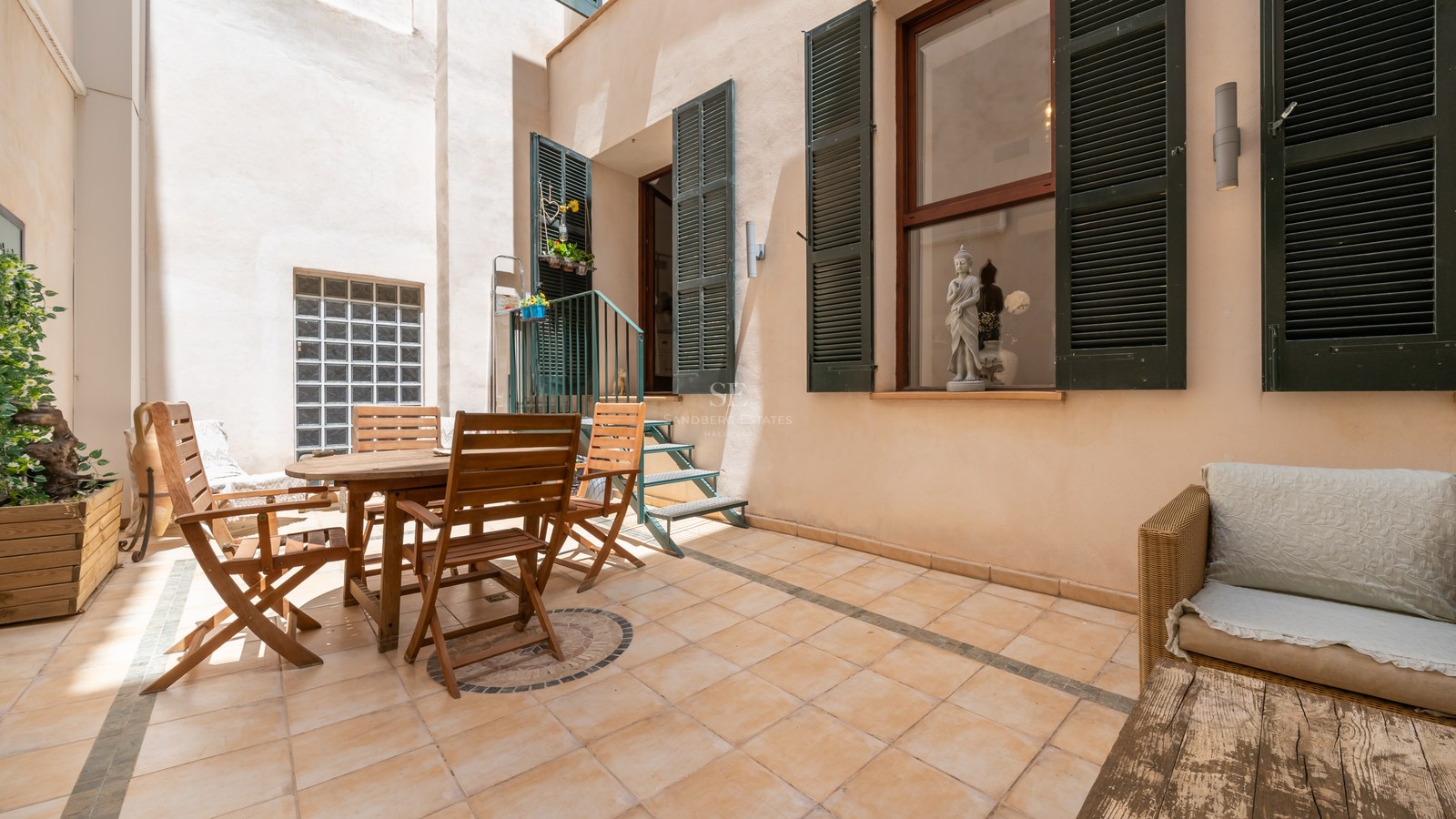 Terrasse privée carrelée avec table à manger en bois, volets verts et salon extérieur en plein jour.
