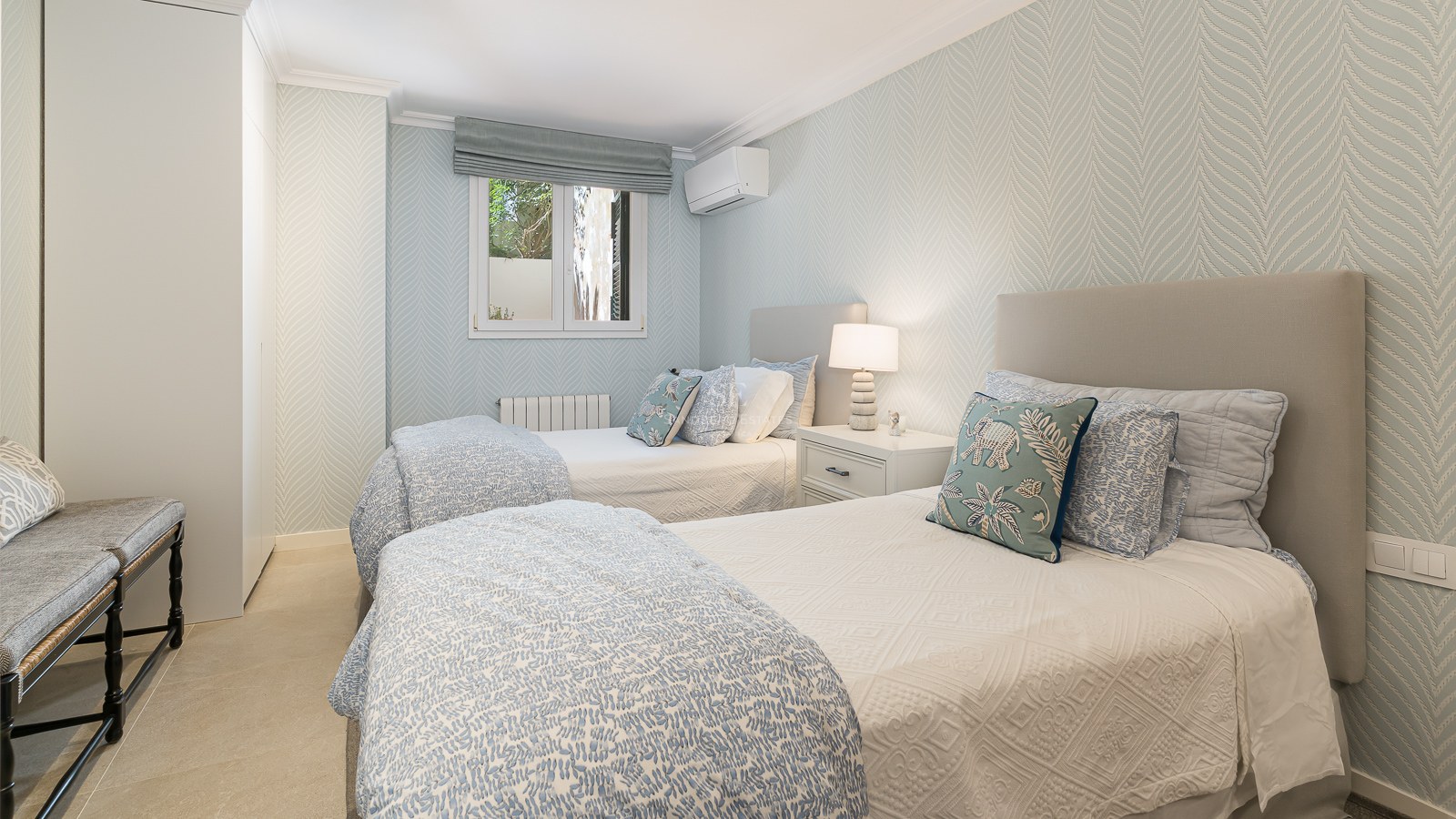 Chambre lumineuse avec deux lits simples, papier peint bleu clair, armoires encastrées et sol en pierre naturelle.