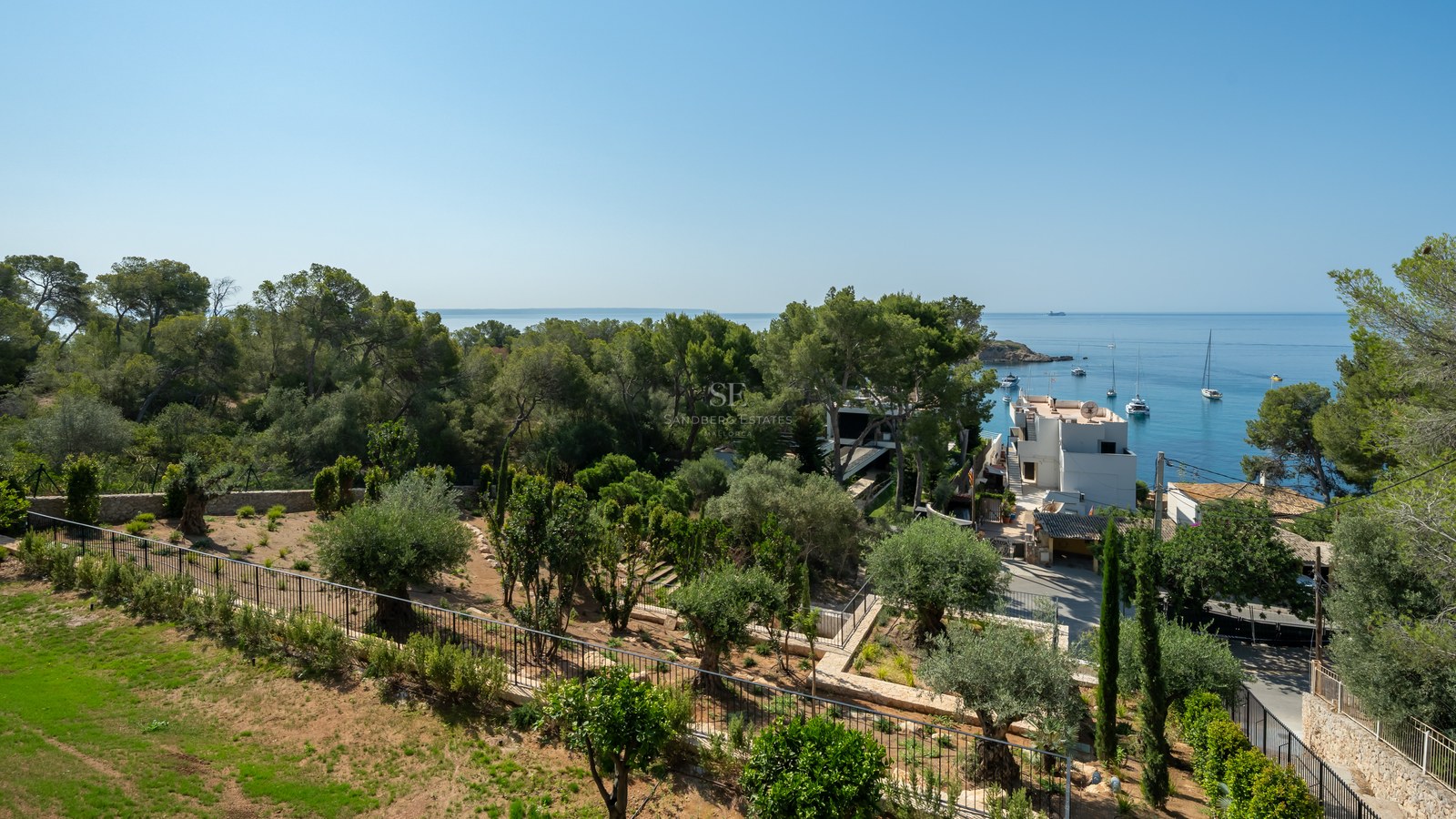 Elevated view of a Mediterranean garden with olive trees overlooking a blue bay with sailboats and white houses.