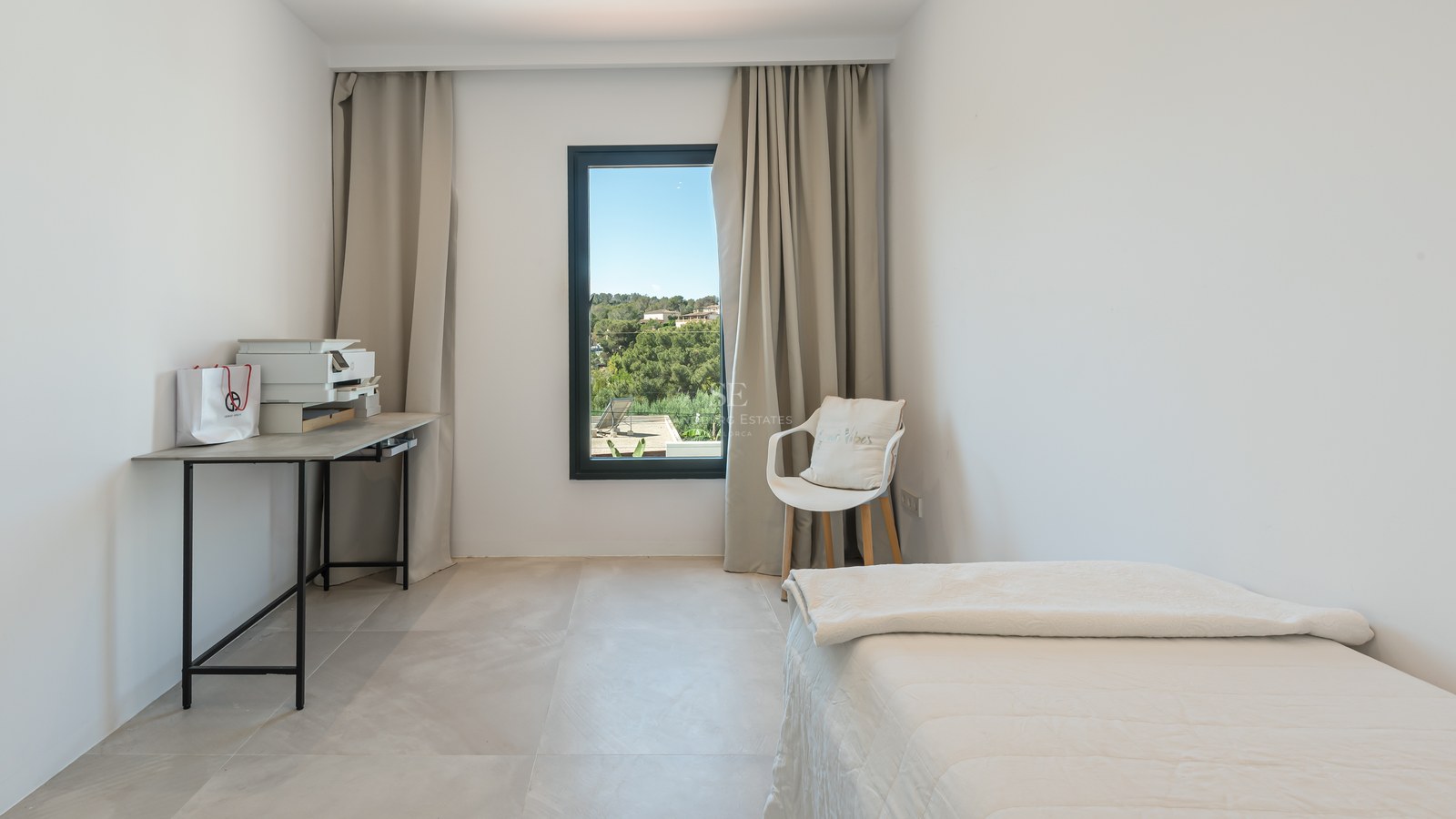 Bright bedroom featuring a white bed, minimalist desk, and a window overlooking green hills.