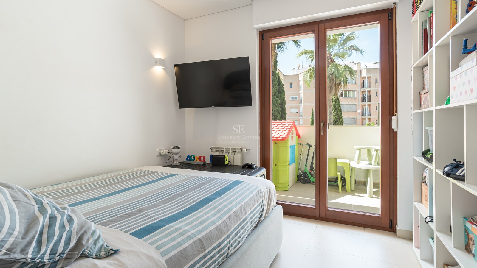Dormitorio luminoso con cama de rayas, TV en pared y puertas de cristal que dan a una terraza con juegos infantiles.