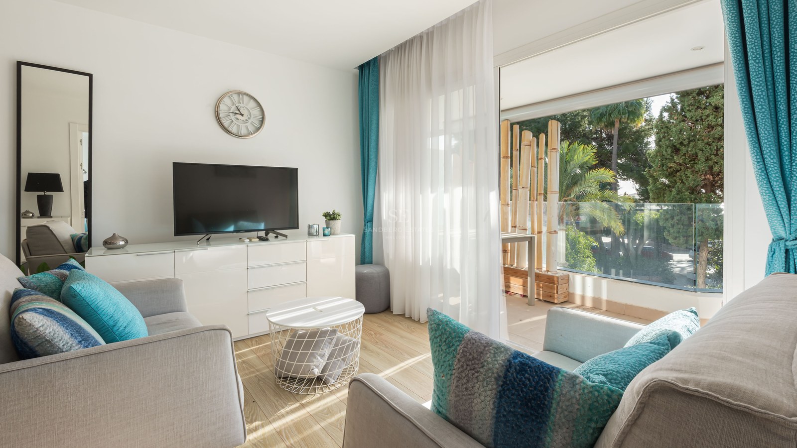 Modern living room with grey sofas, turquoise pillows, and large glass doors opening to a balcony with greenery.