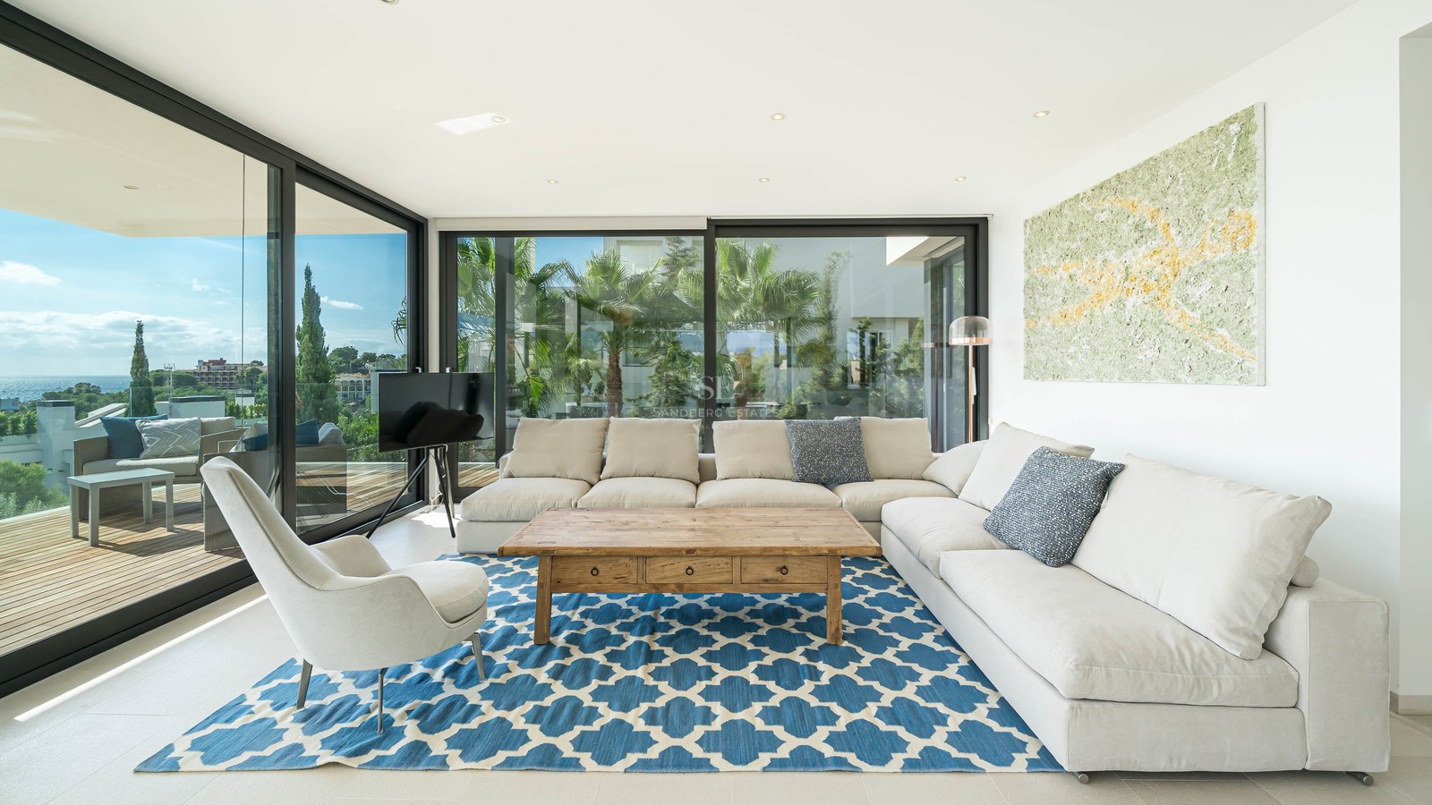 Salon moderne avec canapé en L blanc, tapis bleu à motifs et baies vitrées offrant une vue sur la côte.