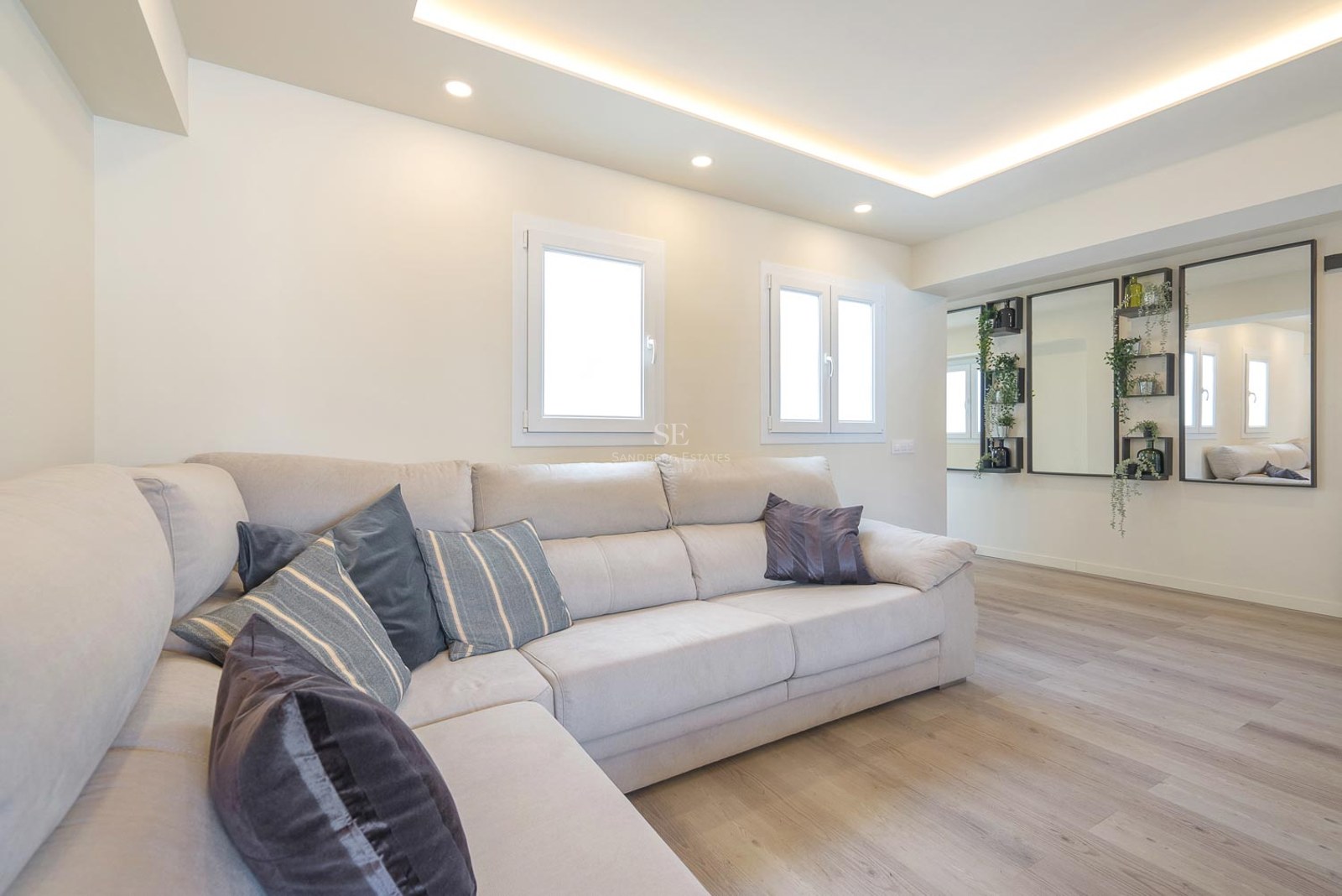 Salon moderne avec un grand canapé en L crème, parquet clair et éclairage au plafond encastré.