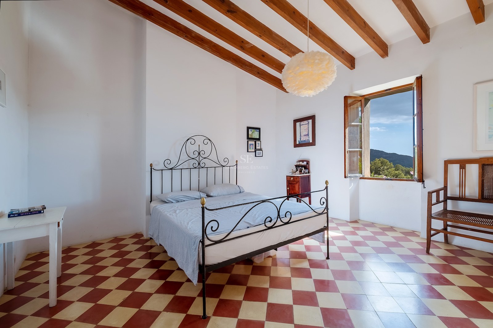 Chambre avec lit en fer forgé, sol en damier rouge et blanc, poutres en bois et fenêtre donnant sur les collines.