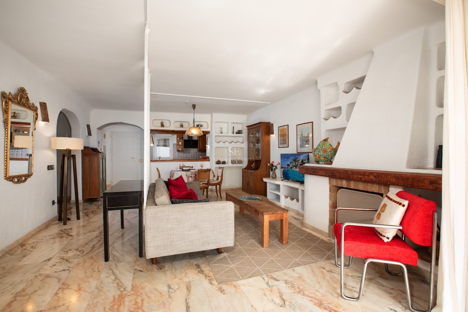 Salon lumineux avec sol en marbre poli, grande cheminée blanche, canapé gris et fauteuil rouge.