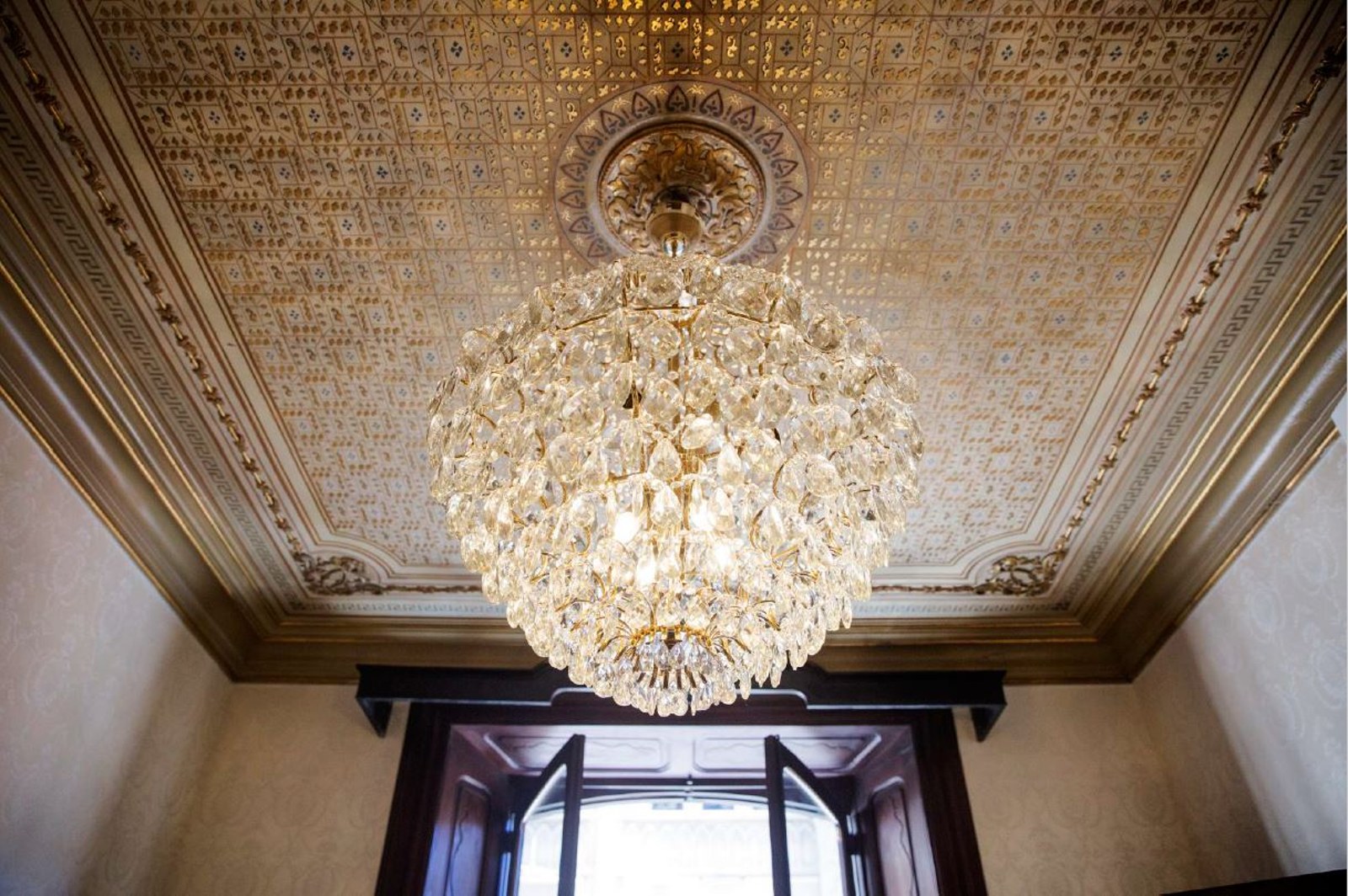 A large crystal chandelier hanging from a highly decorative gold-patterned ceiling with ornate moldings.