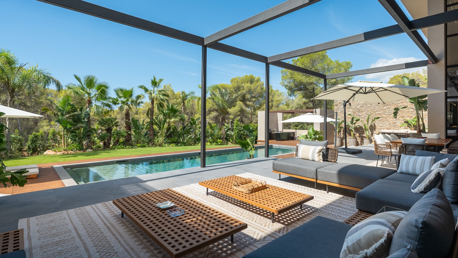 Terrasse moderne avec canapés gris, tables en bois et piscine face à un jardin tropical luxuriant.