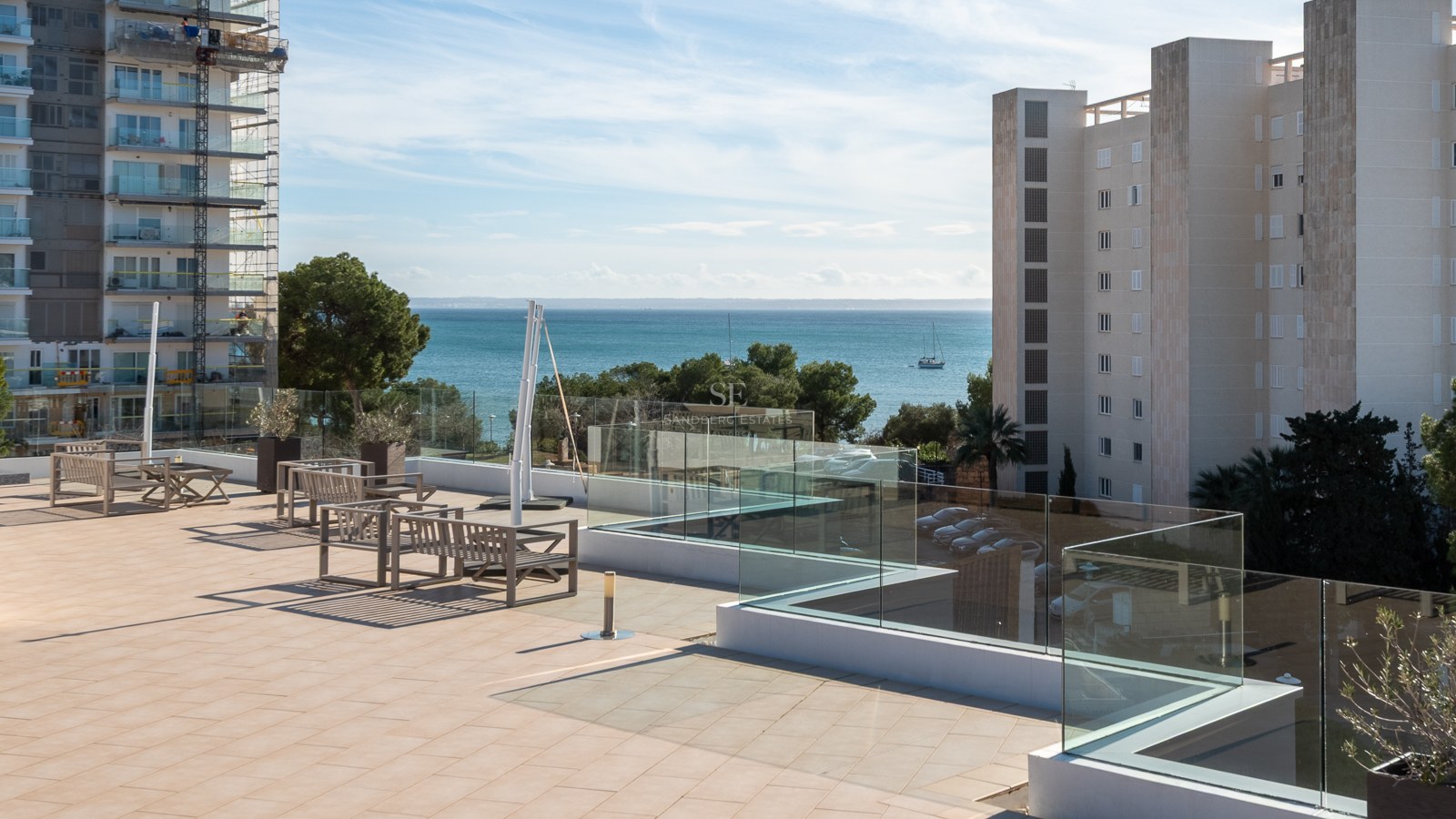 Spacious tiled terrace with glass railings and wooden seating, overlooking the blue sea and coastal buildings.