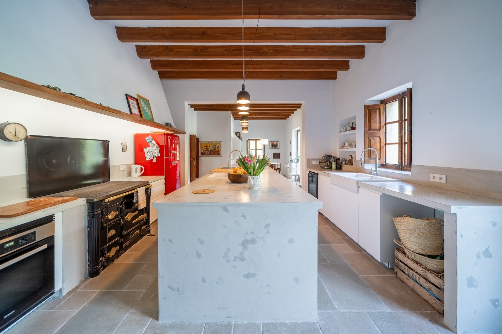 Grande cuisine avec îlot central blanc, poutres en bois apparentes, réfrigérateur rouge rétro et piano de cuisson.