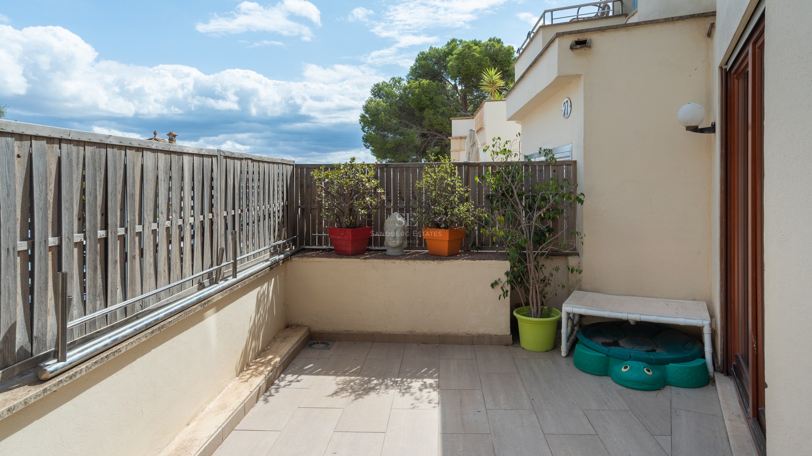 Terraza privada con suelo cerámico, valla de madera, plantas en macetas y vistas a los árboles bajo un cielo azul.