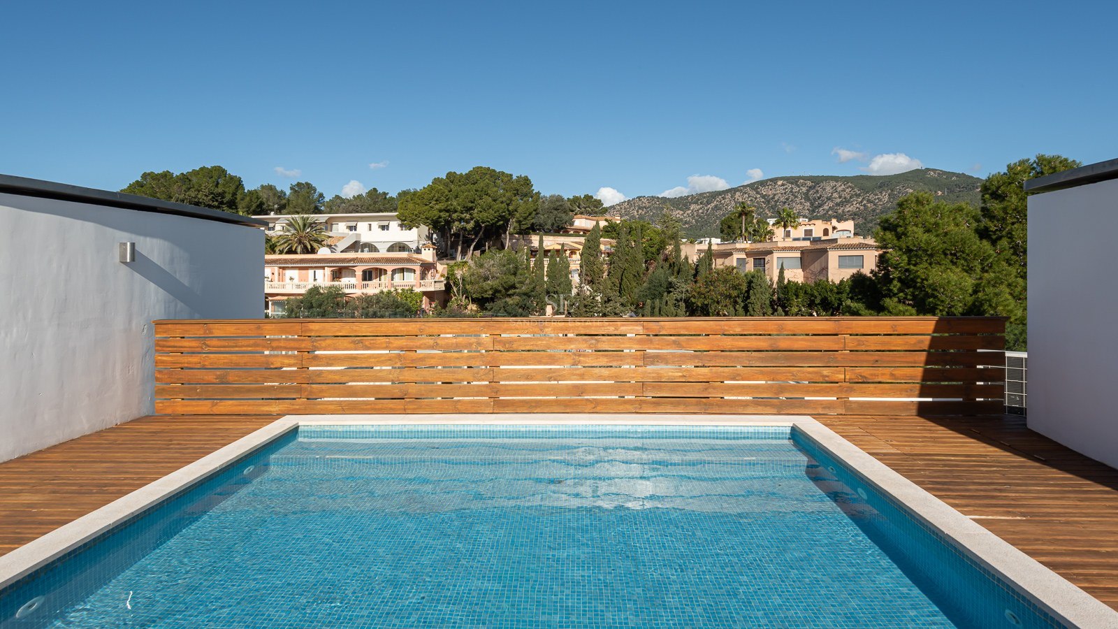 A rectangular rooftop swimming pool with blue tiles and a wooden deck, overlooking green hills and houses.