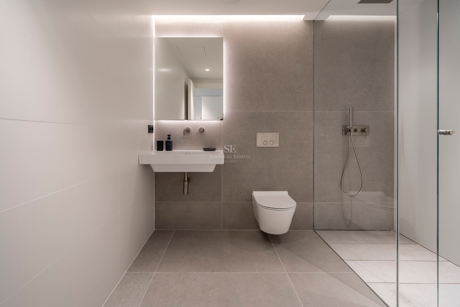Salle de bain moderne avec carrelage gris, miroir rétroéclairé, lavabo blanc, WC suspendu et paroi de douche en verre.